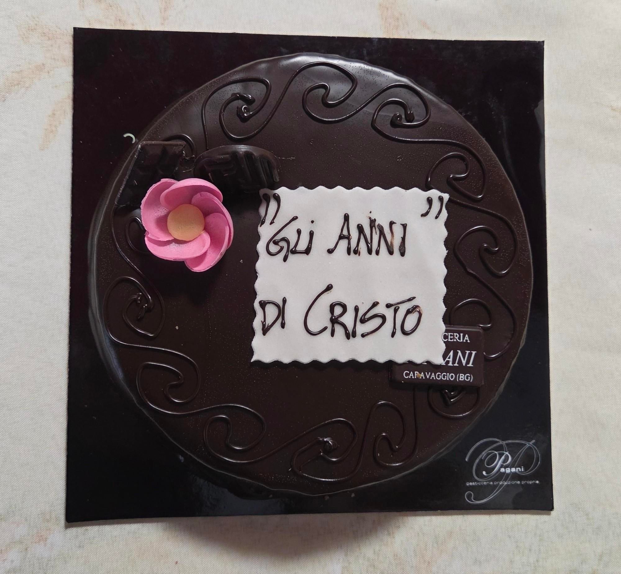 Torta al cioccolato tonda con targhetta bianca con scritto sopra "gli anni di cristo" e fiorellino rosa 
