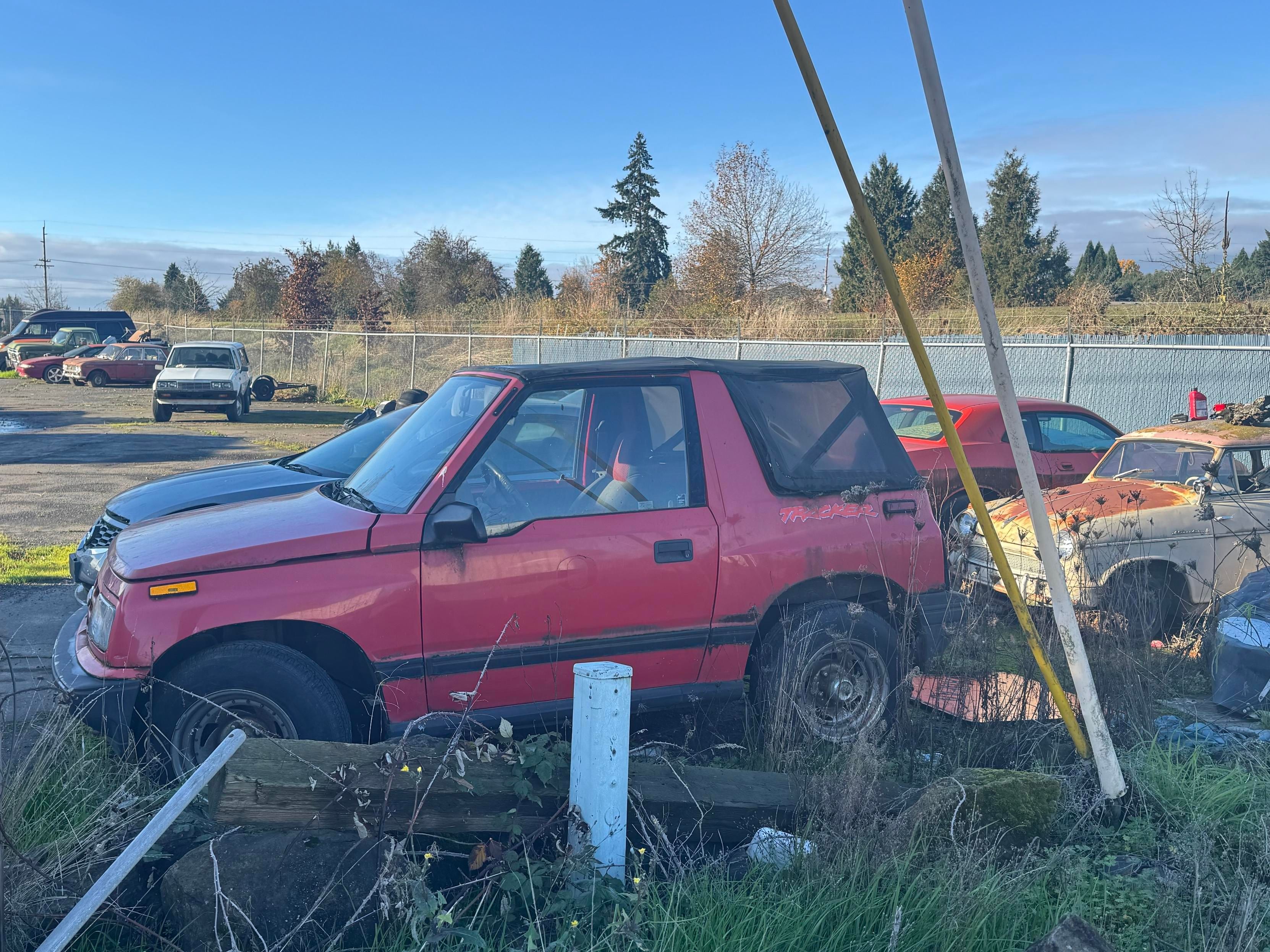 A red, weathered SUV is parked in a junkyard, surrounded by various old and rusty vehicles. Overgrown grass and weeds are visible, and the background includes a chain-link fence and scattered trees. The sky is blue with some clouds.