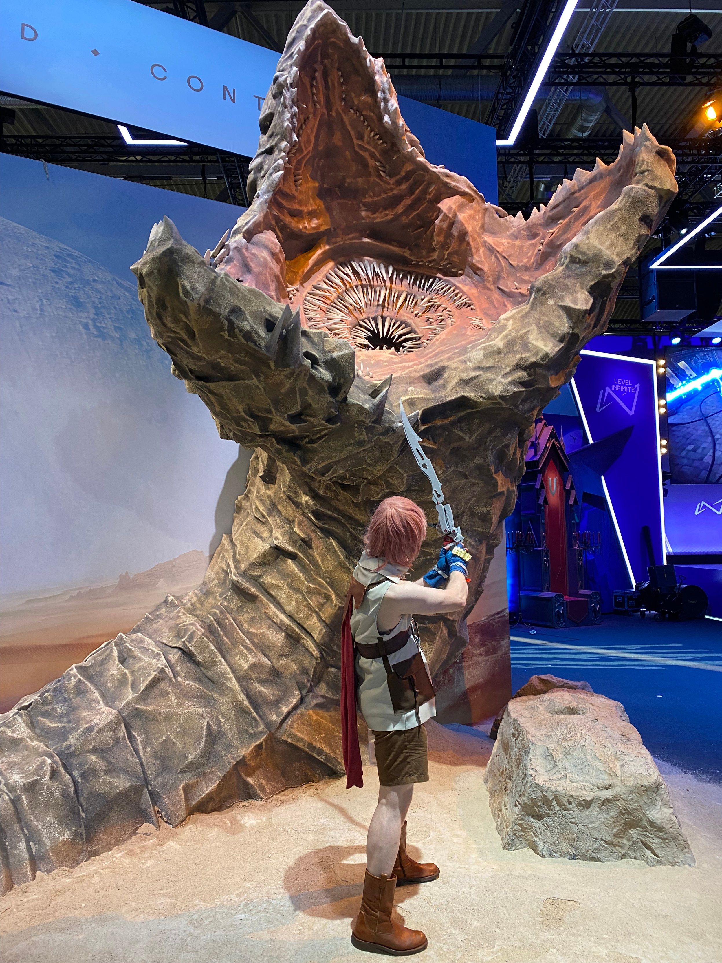 Me in my Lightning cosplay at Gamescom, in front of a model of a giant sandworm from Dune. I look at the worm, face turned away from the camera, pointing the gunblade at the worm
