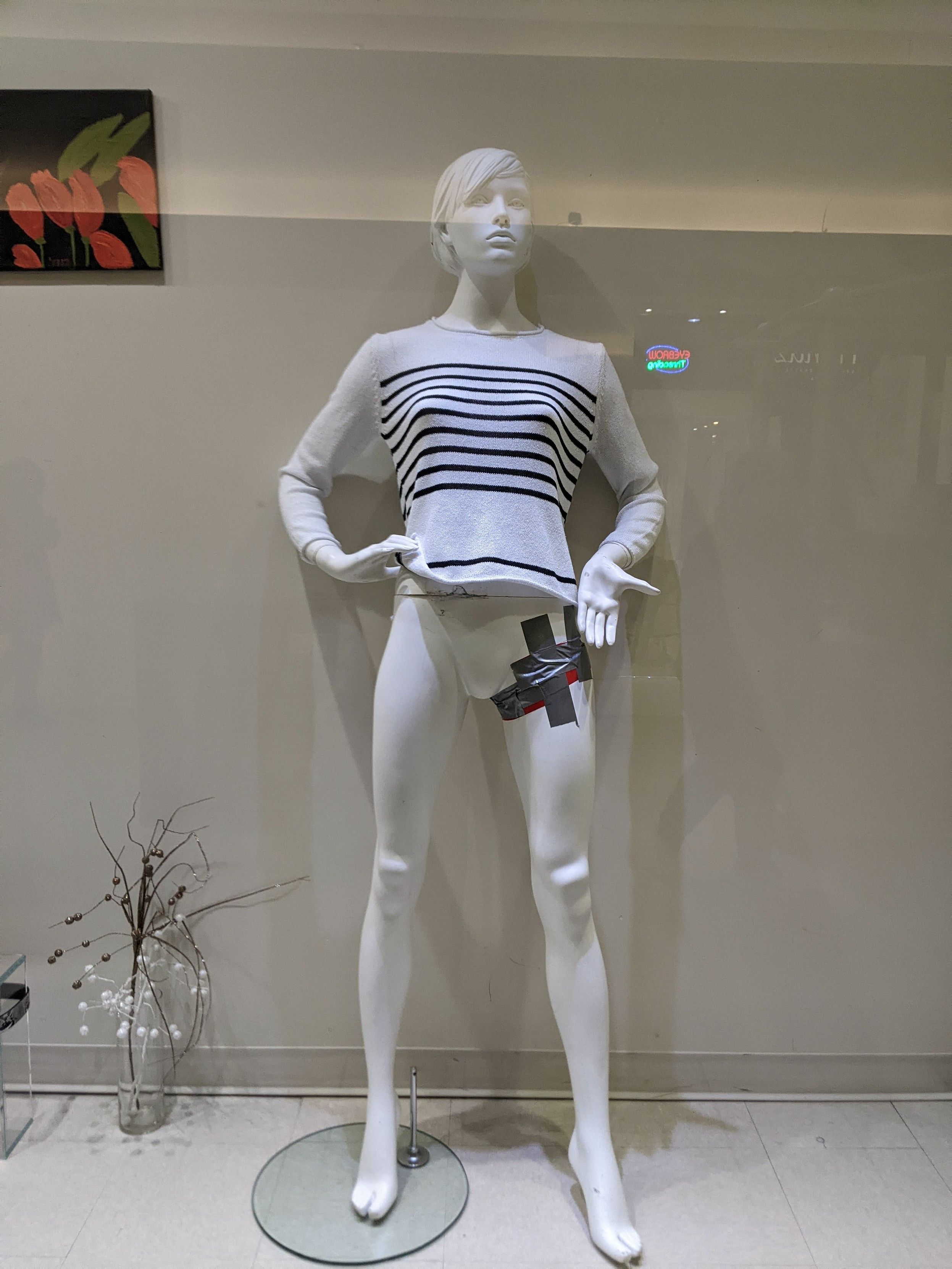 A mannequin wearing a white and black striped shirt and no pants. Its leg/groin area has been secured with duct tape. Its palm is facing the viewer, thumb out, as if to say "check out my taped up limb". 