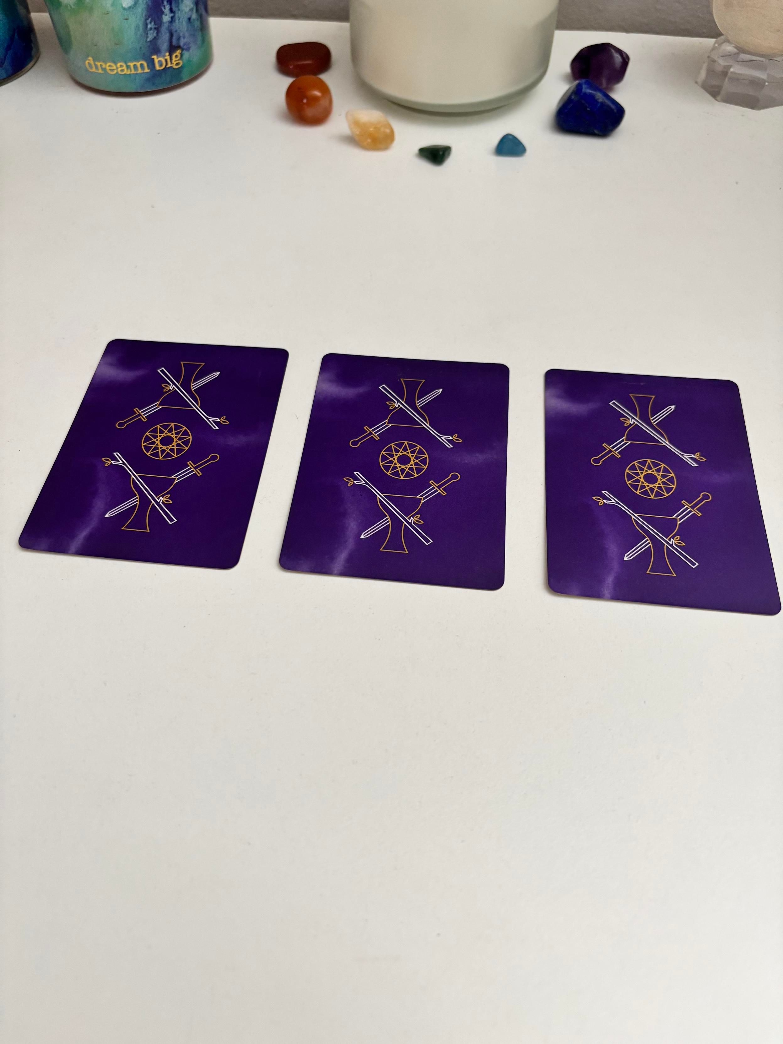 three tarot cards face down on a white table. they have purple backs with a gold and white design featuring two pentacles overlapping to make a ten pointed star in the center, and on either side of that there is a cup with a wand and a sword crossing it. also on the table are a white candle in a jar with stones of different rainbow colors surrounding it, and a blue and green candle holder with gold words that say dream big. 