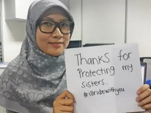 A woman, wearing glasses and a hijab, holds up a hand-written sign that reads "Thanks for protecting my sisters. #illridewithyou"