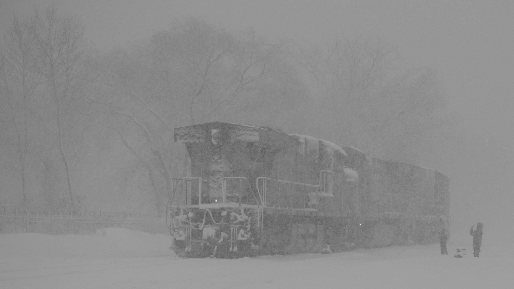 A pair of diesel locomotives moving slowly through a near-whiteout blizzard. Two children, ankle deep in snow, wave to it.