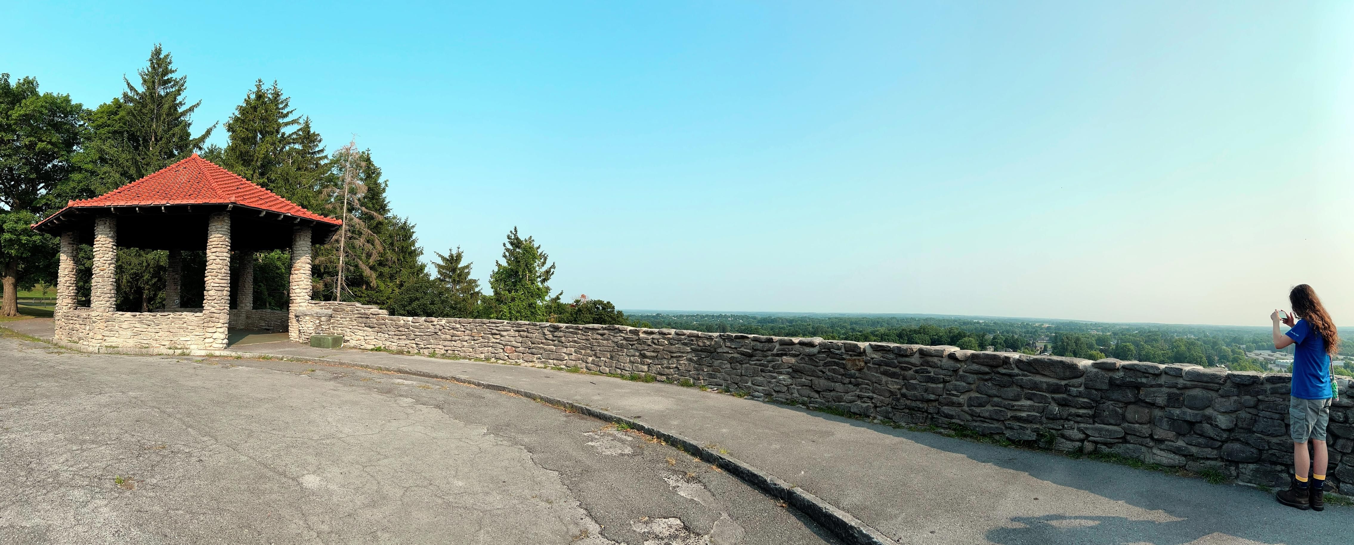 A semi-panoramic photo of the lookout at Thompson Park. The sky above is cloudless and shades from turquoise in the top left to a pale off white on the far right. On the left is a sort of gazebo built of stones, with pillars supporting a sienna orange roof. Trees paint a green border between the structure and the sky. From there a low stone wall stretches across the picture until on the far left there is a long-haired person in blue T-shirt, cargo shorts, and hiking boots, holding a phone and taking a picture of the view.