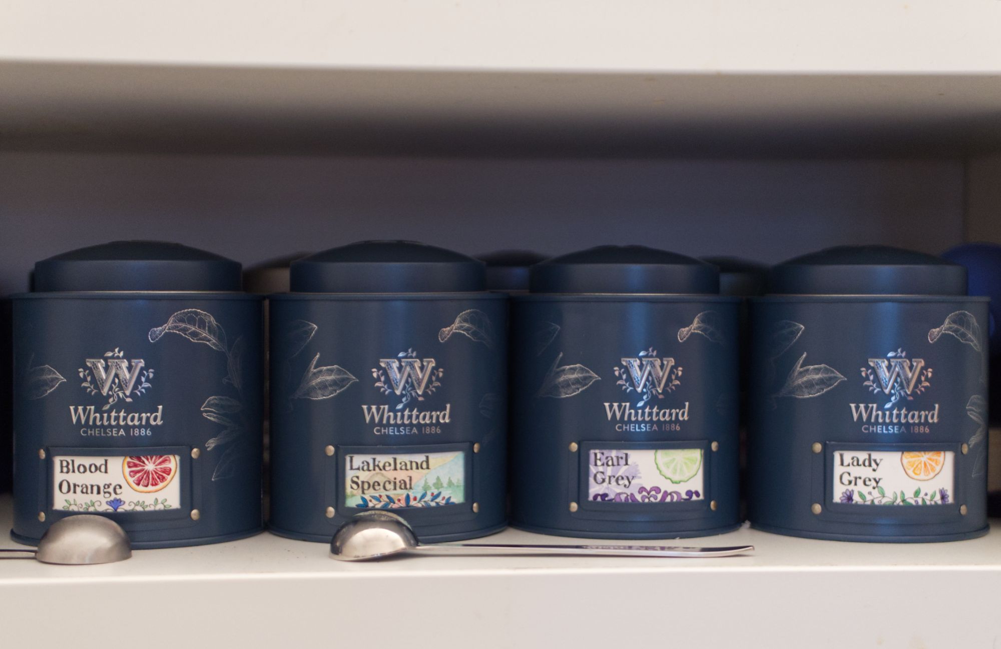 4 More stout teal tea caddies with labels that read: Blood Orange, Lakeland Special, Earl Grey, and Lady Grey. 