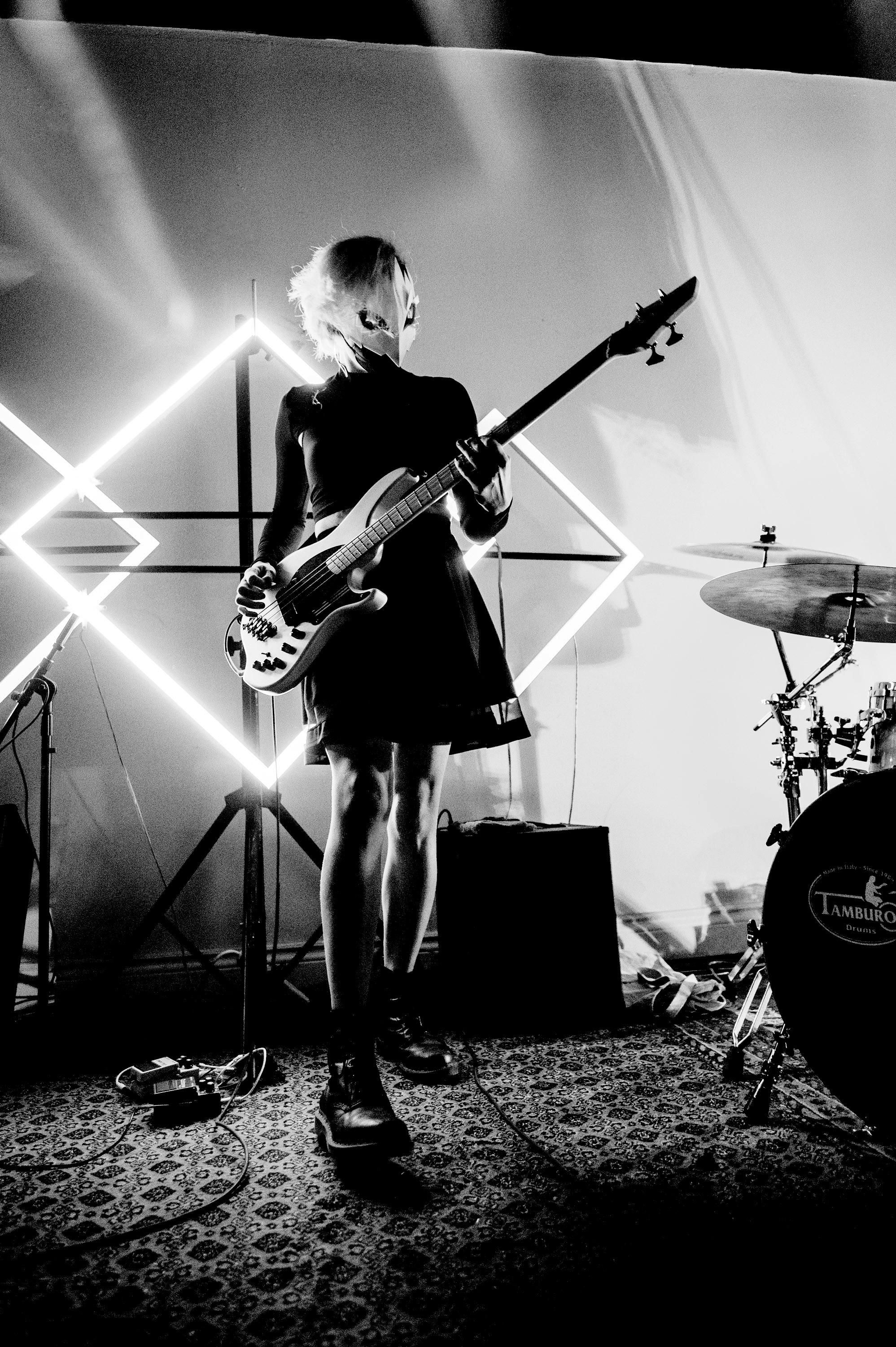 A girl in a black dress standing in front of diamond-shaped LED lights on a stage, playing bass guitar. Photo by Alex Carre
