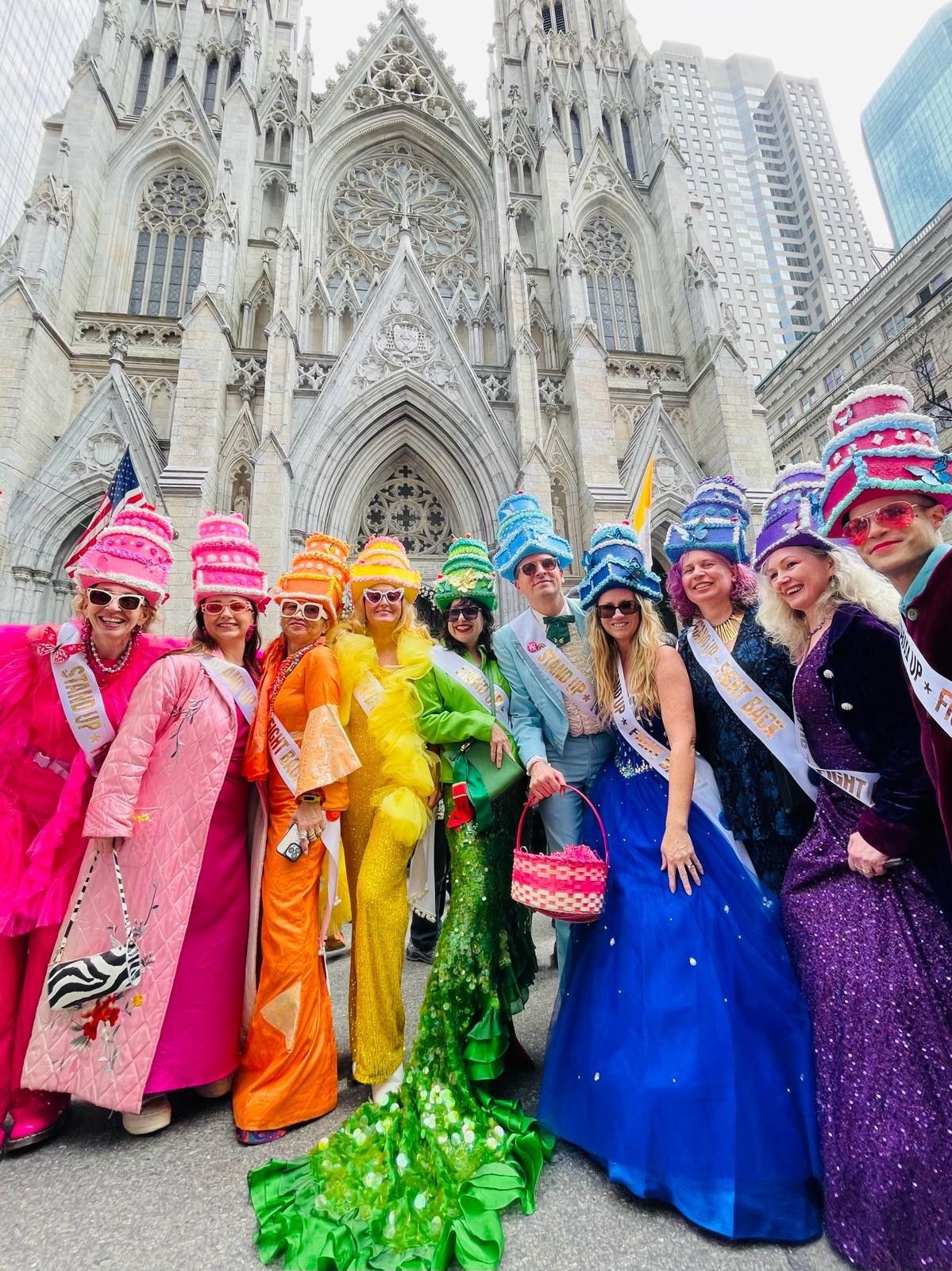 People including me wearing 3 tiered cake hats and costumes in a rainbow of colors in front of St Patrick's Cathedral in NYC