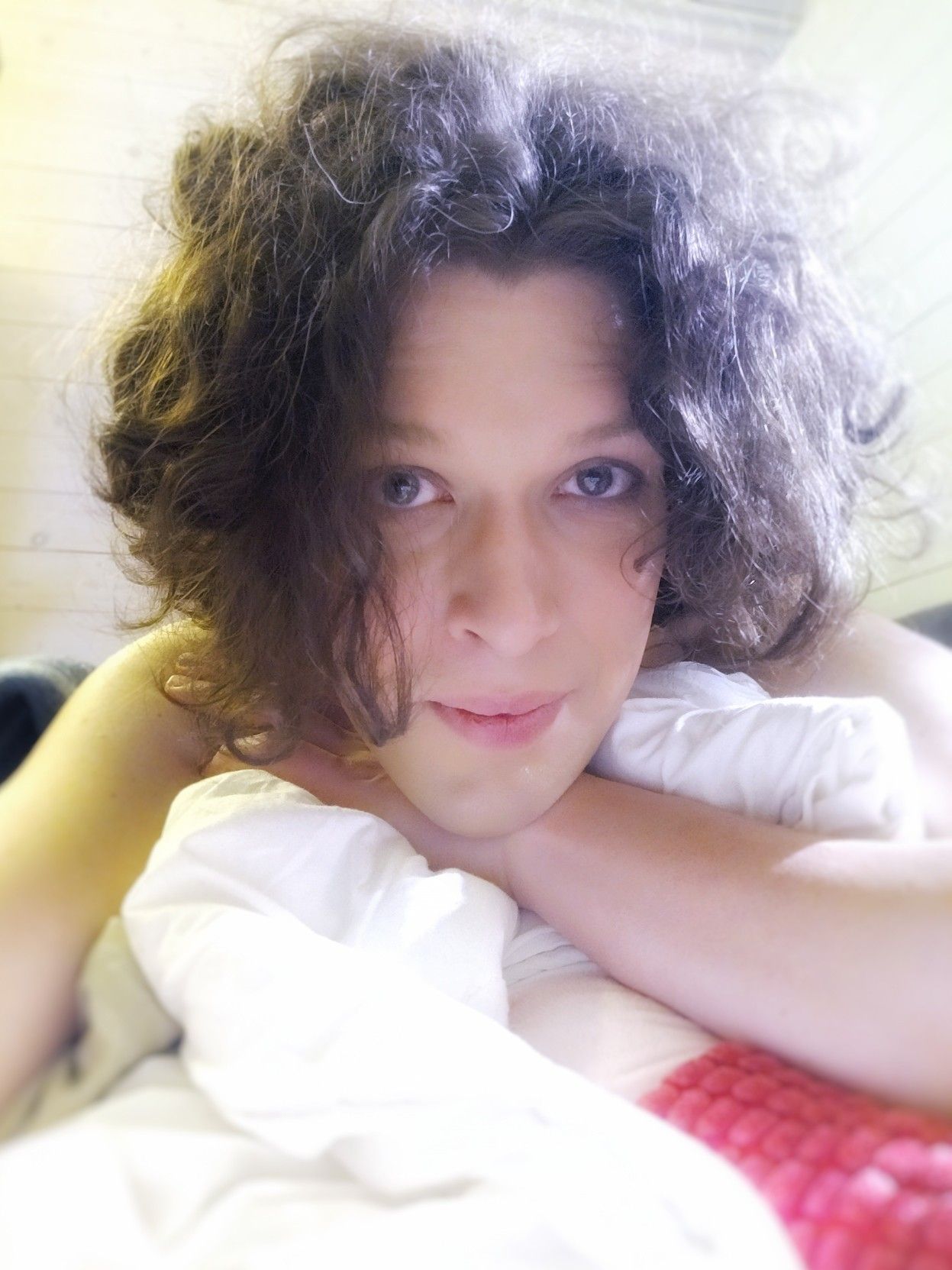 Selfie from in front, me lying on my belly on my bed, resting me head on my left wrist on a bunched up, white blanket, looking into the camera and smiling with a patient anticipation, sunlight from the window catching in my messy hair.