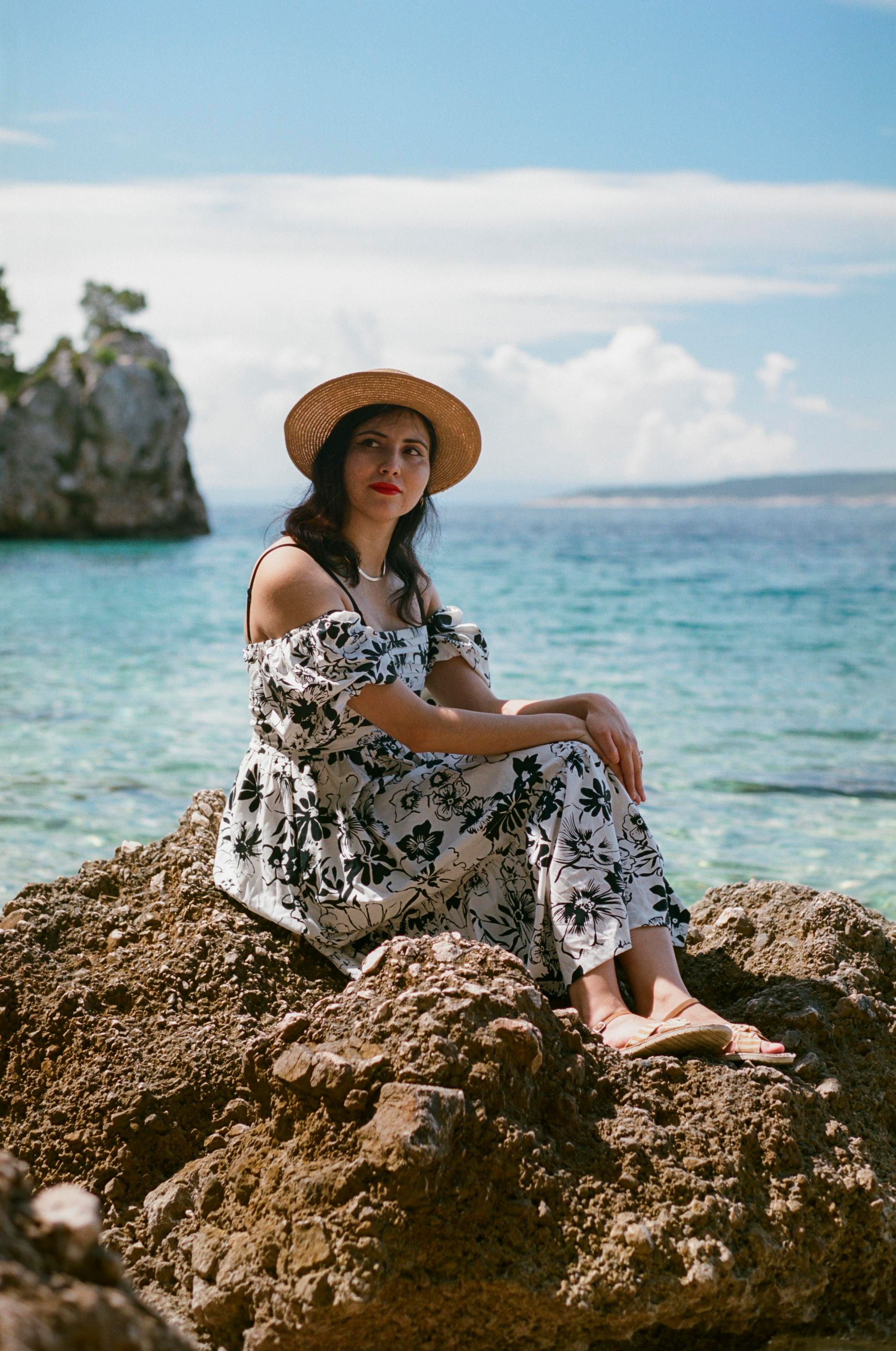 A beautiful woman with dark hair, wearing a gold necklace, a straw hat, and a long white and black summer dress, sitting on a rock in front of the Mediterranian sea.