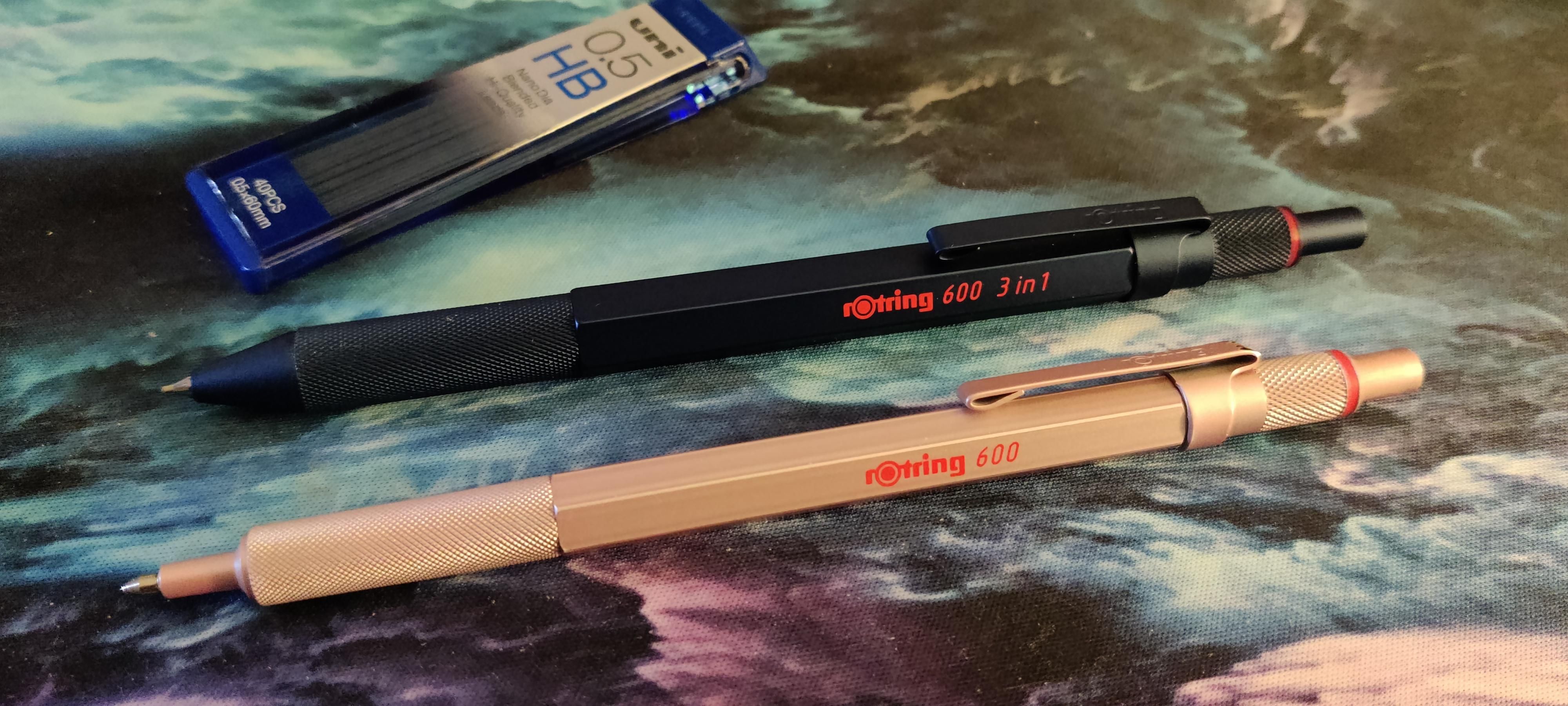 A pink Rotring 600 pen and a dark blue Rotring 600 3-in-1 pen. Both have a knurled grip and hexagonal barrels, a thin metal pocket clip, and the "Rotring 600" printed in bright red on the side. A container of Uni Nano Dia 0.5mm HB mechanical pencil lead is sitting in the background.