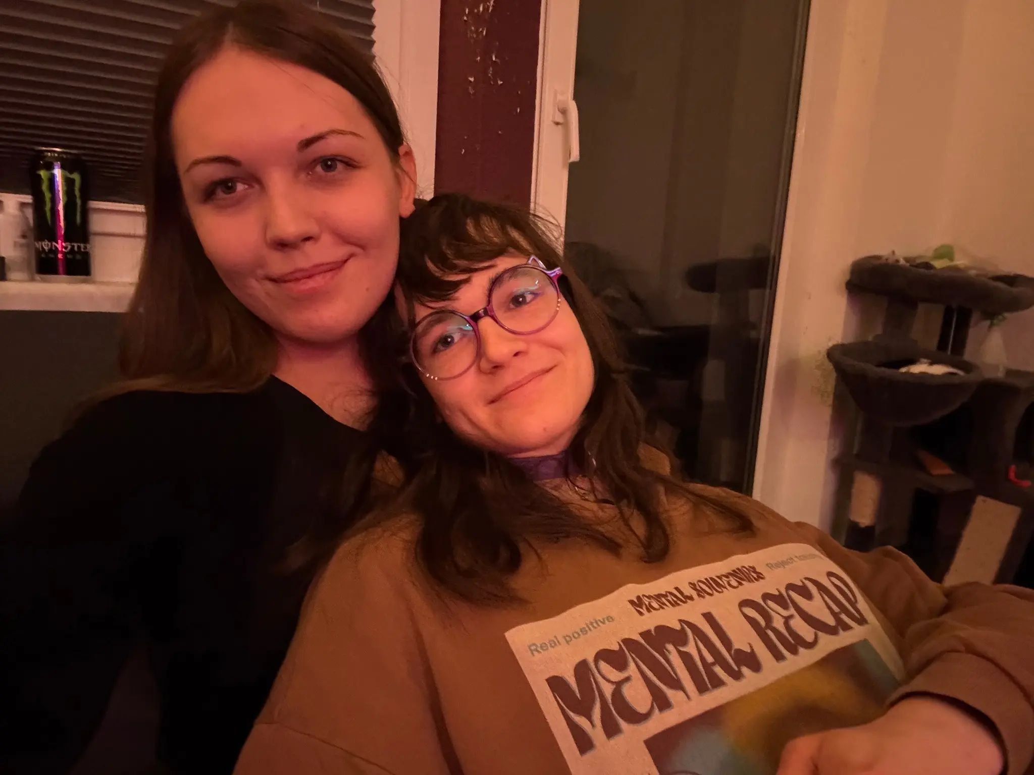 Photo of two feminine people taking a selfie together indoors. The woman on the left has long, straight dark brown hair, defined eyebrows, and wears a black top. The person on the right has wavy, shoulder-length dark brown hair with lighter highlights, wears large round purple-framed glasses with cat ears on the frame. Both are cuddling with each other and looking at the camera, smiling.