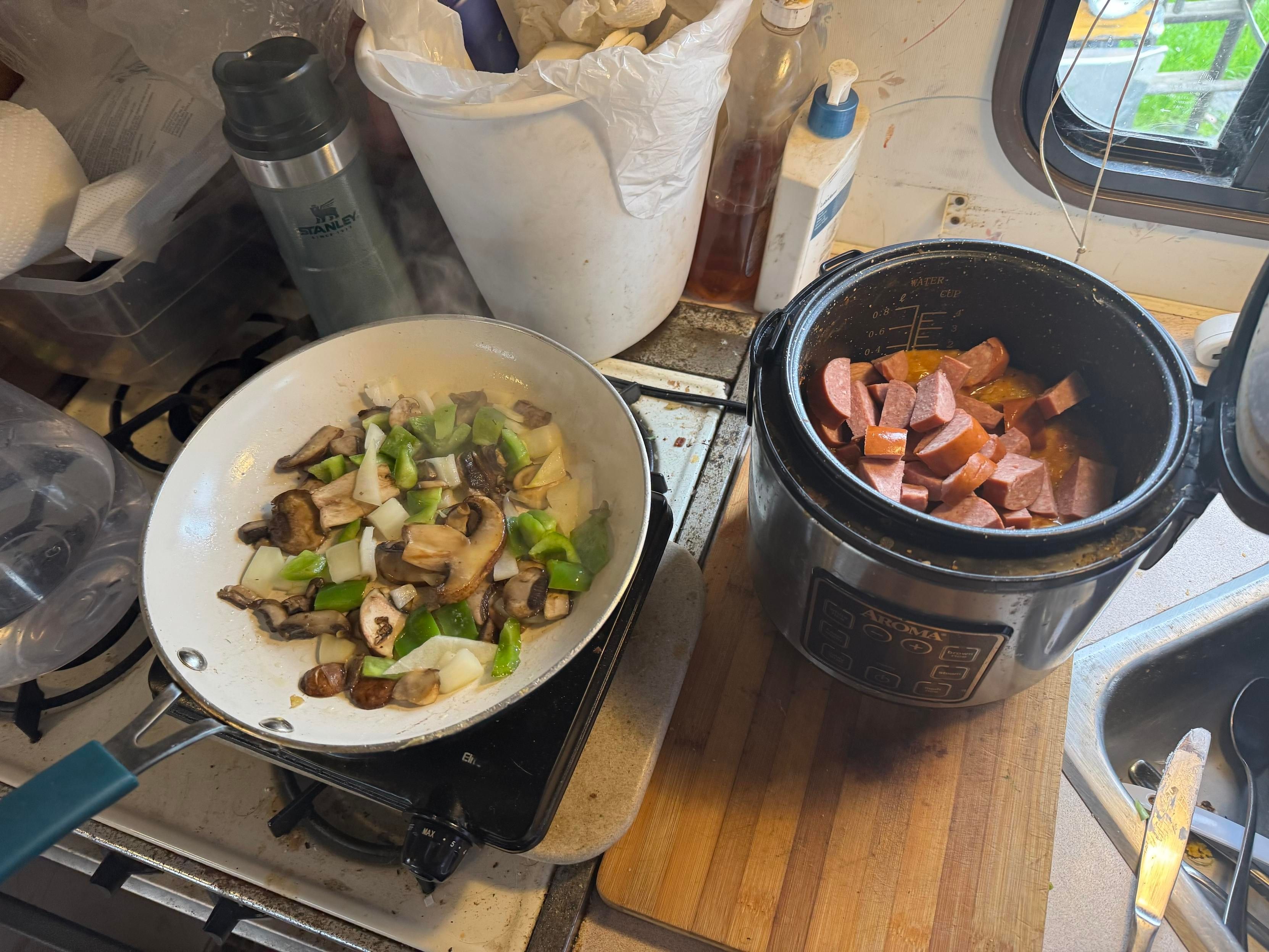 A pan on the stove with sautéing mushrooms, green peppers, and onions, next to a slow cooker containing sliced sausage. The countertop is cluttered with a thermos, bottles, and other kitchen items.