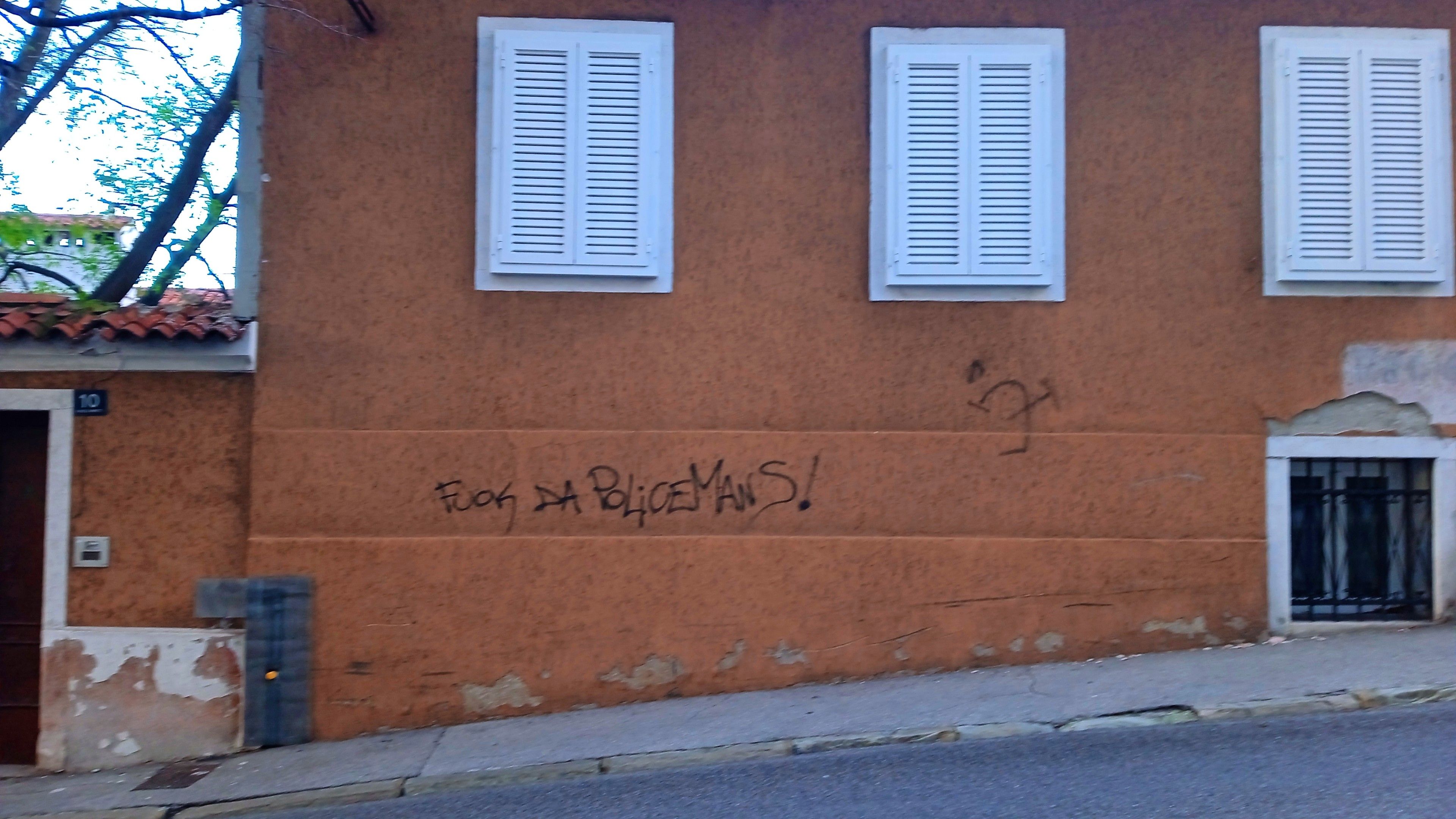 A black writing on an orange-brown wall with tree windows closed by white shutters that says "fuck da policemans" and a distorted  and completely wrong hammer and sickle symbol with a small star near it