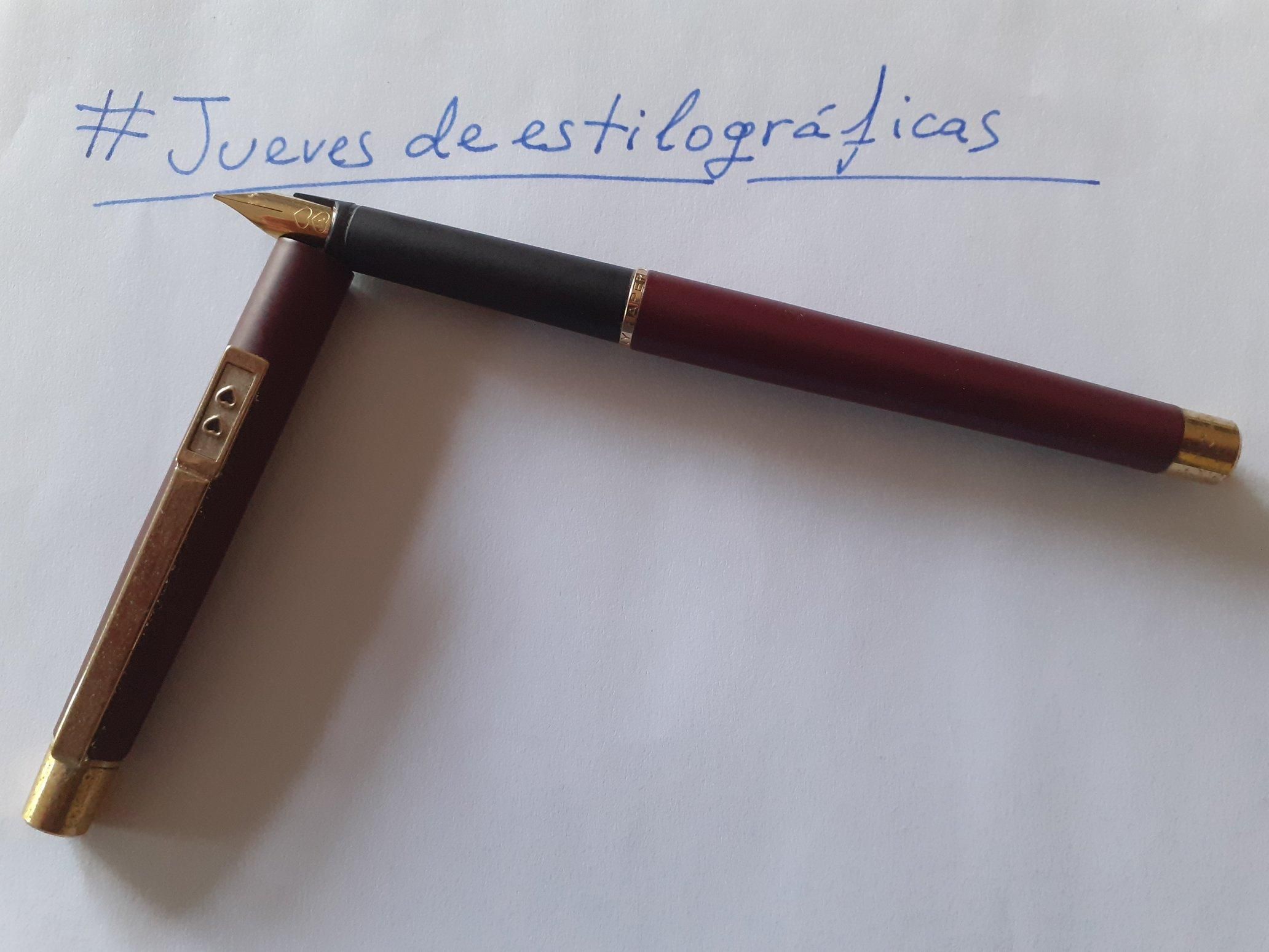 La imagen muestra una pluma estilográfica colocada en diagonal sobre un papel blanco. La pluma es de color granate y negro, con detalles dorados. El plumín es dorado. Junto a ella está el capuchón, también granate. El el clip de sujección, de color dorado, se ven los dos corazones de la marca Paper Mate. En el papel se ha escrito la frase "jueves de estilográficas" en tinta azul.