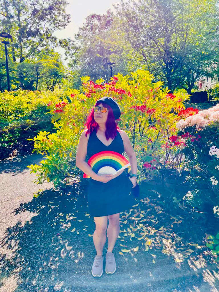 Crissy wearing black top and skirt, standing outside, displaying pride rainbow hand fan, behind her are plants with green leaves and red flowers 