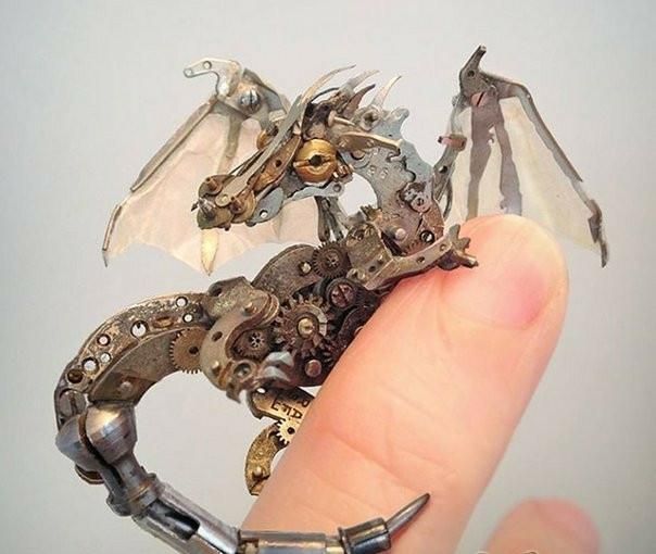 Tiny sculpture in the shape of a winged drsgon resting on the fingertip of a white hand and created with tiny cogs and other parts of watches