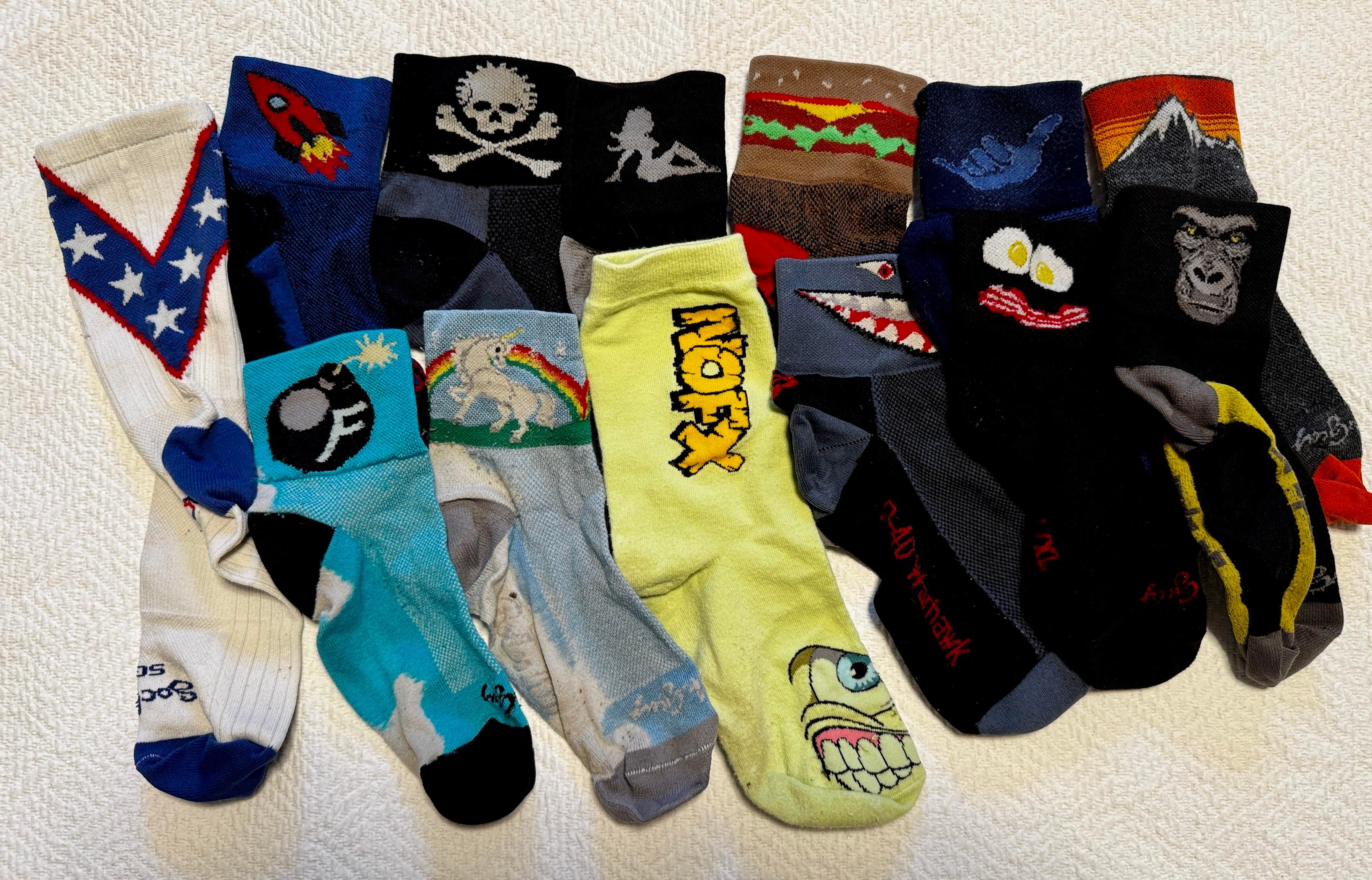A photo of various whimsical socks, including Evel Knievel, rockets, F-bombs, skull and cross bones, rainbow and unicorns, hauling ass ( naked lady silhouette on mud flaps), NoFX, hamburger, P-51 mustang shark, hang loose hand signal, bacon and eggs in a smiley face, a mountain top, and a gorilla.