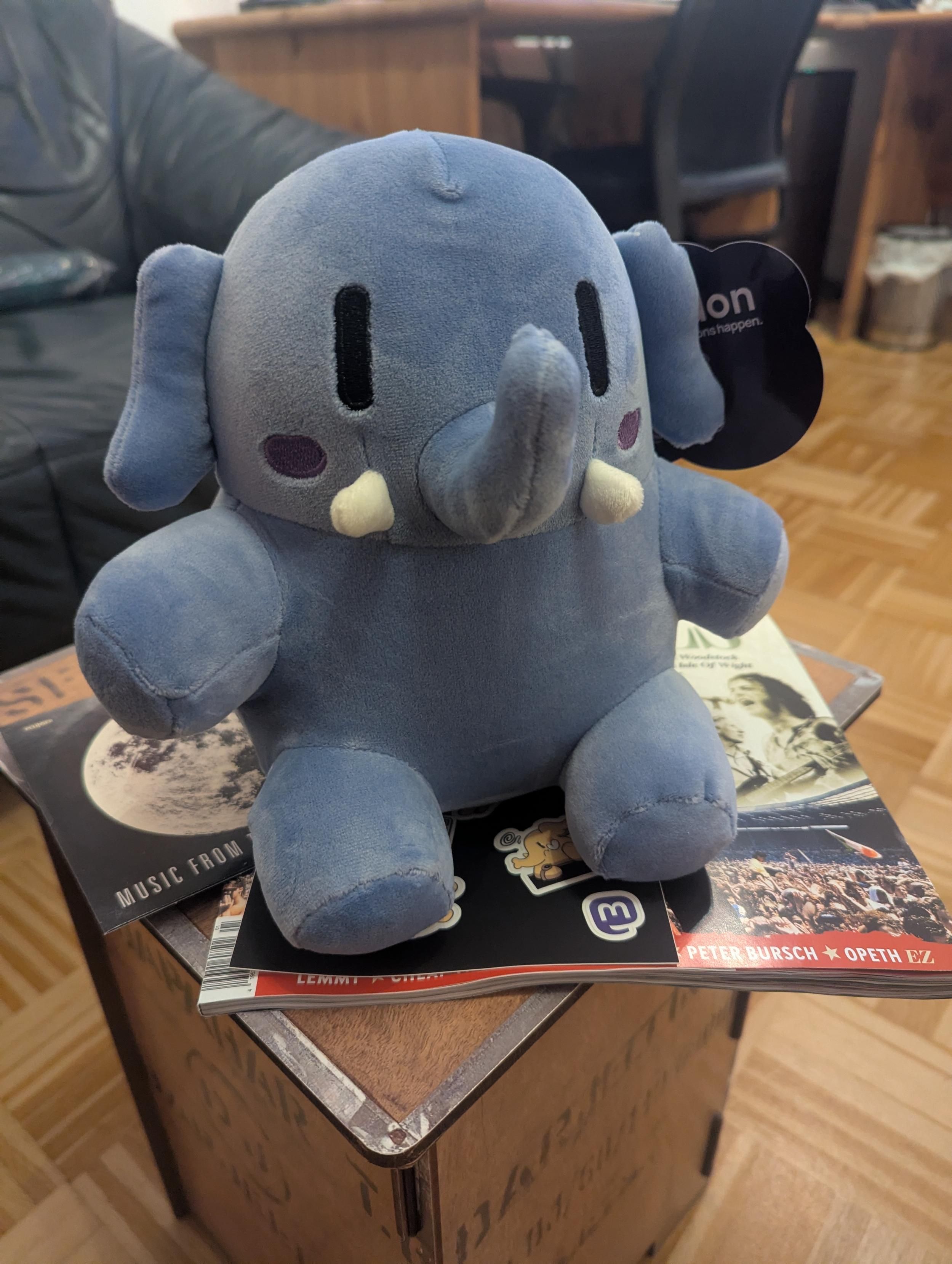 Mini Mastodon plushie Blue sitting on a small wooden stool in the living room. There's the most recent eclipsed magazine sticking out on the side, so it's more specifically sitting on that.