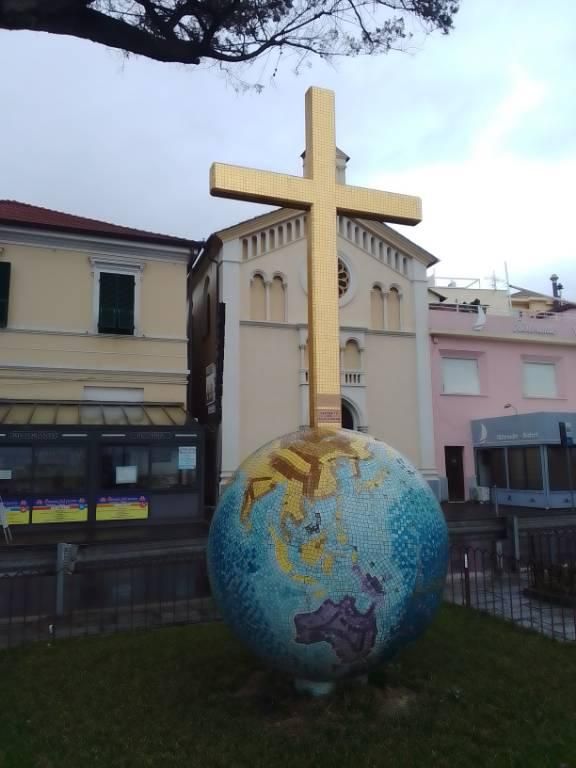 Una foto a colori, in formato verticale, che ritrae un pratino quadrato circondato da un cancelletto basso al centro del quale c'è una scultura, un globo terrestre il cui diametro sarà di circa 2 metri e la cui superficie, con i mari e i continenti, è un mosaico di diversi colori. Dalla sommità del globo si innalza una grossa croce da tortura, mosaicata di tessere dorate, altra circa 3 metri. Sullo sfondo c'è una strada e dietro quella la facciata di una chiesa tra due case basse. Sopra c'è il cielo un po' grigio e nuvoloso e un ramo di un pino che sbuca dall'angolo sinistro in alto dell'inquadratura.