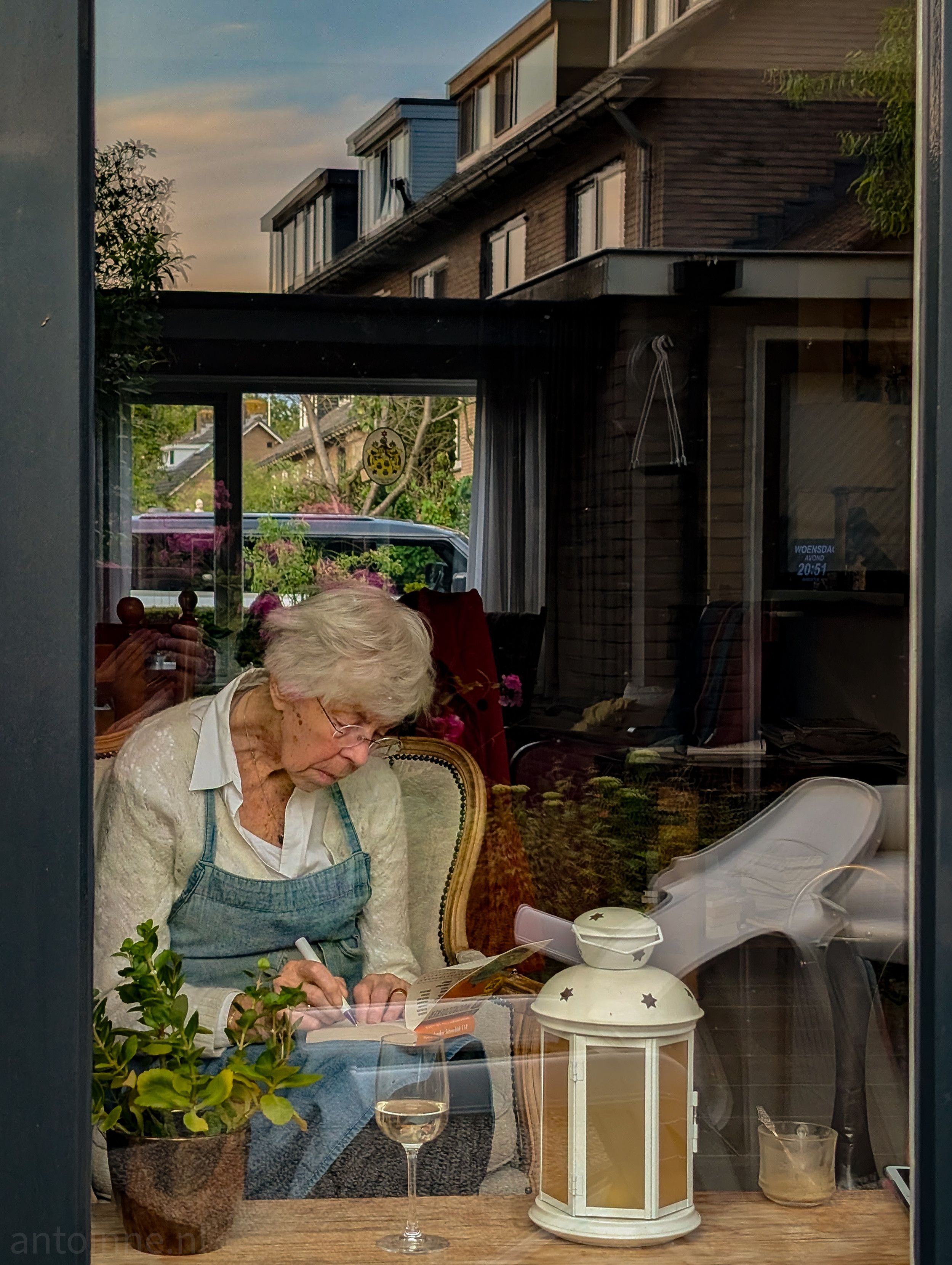 An elderly person with short white hair is sitting in a chair, seen through a window. She is wearing glasses, a white shirt, and a light blue apron. They are leaning forward, working on a crossword puzzle.

A wine glass is on a wooden surface in front of her, and a white lantern with star cutouts is next to it. Potted plants and other objects are visible, both inside the room and reflected in the window. The background outside the window shows residential buildings and trees under a colorful sky.
