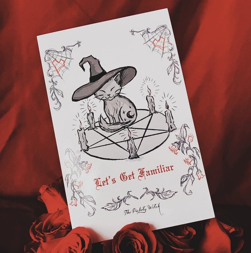 An illustration of a small, smiling cat wearing a witch hat, sitting in the center of a pentagram surrounded by five lit candles. The card features Gothic-style text that reads "Let's Get Familiar" and "The Pickety Witch," decorated with spiderwebs, hearts, and floral vines. The card is resting on a background of red roses and fabric.