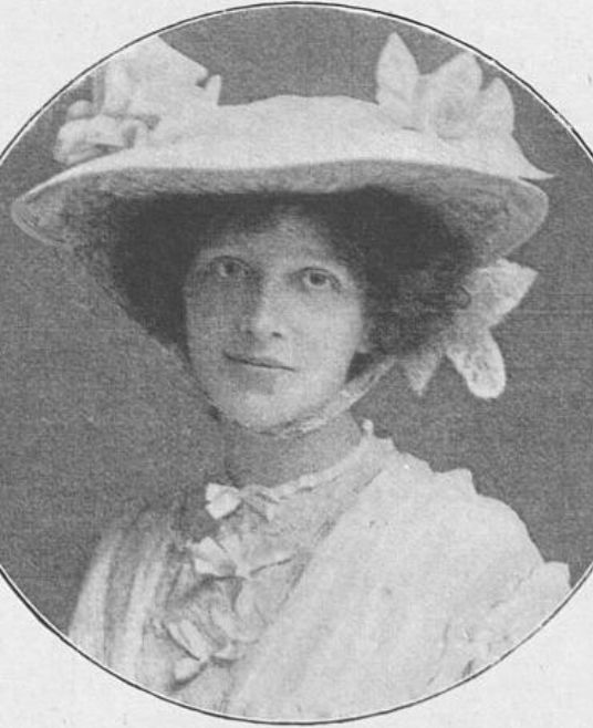 Edith Ostlere, a white woman with dark curly hair, wearing a white hat with a wide brim, and a white dress with lace and bows as trim