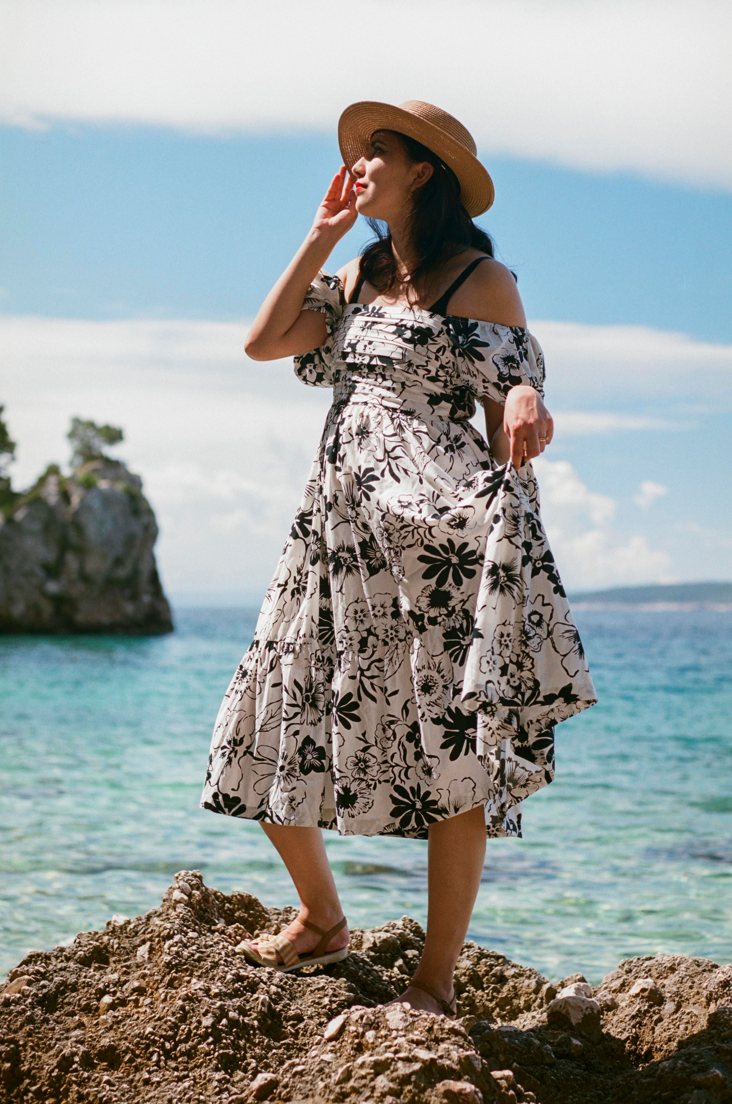 A beautiful woman with dark hair, wearing a gold necklace, a straw hat, and a long white and black summer dress, standing on a rock in front of the Mediterranian sea.