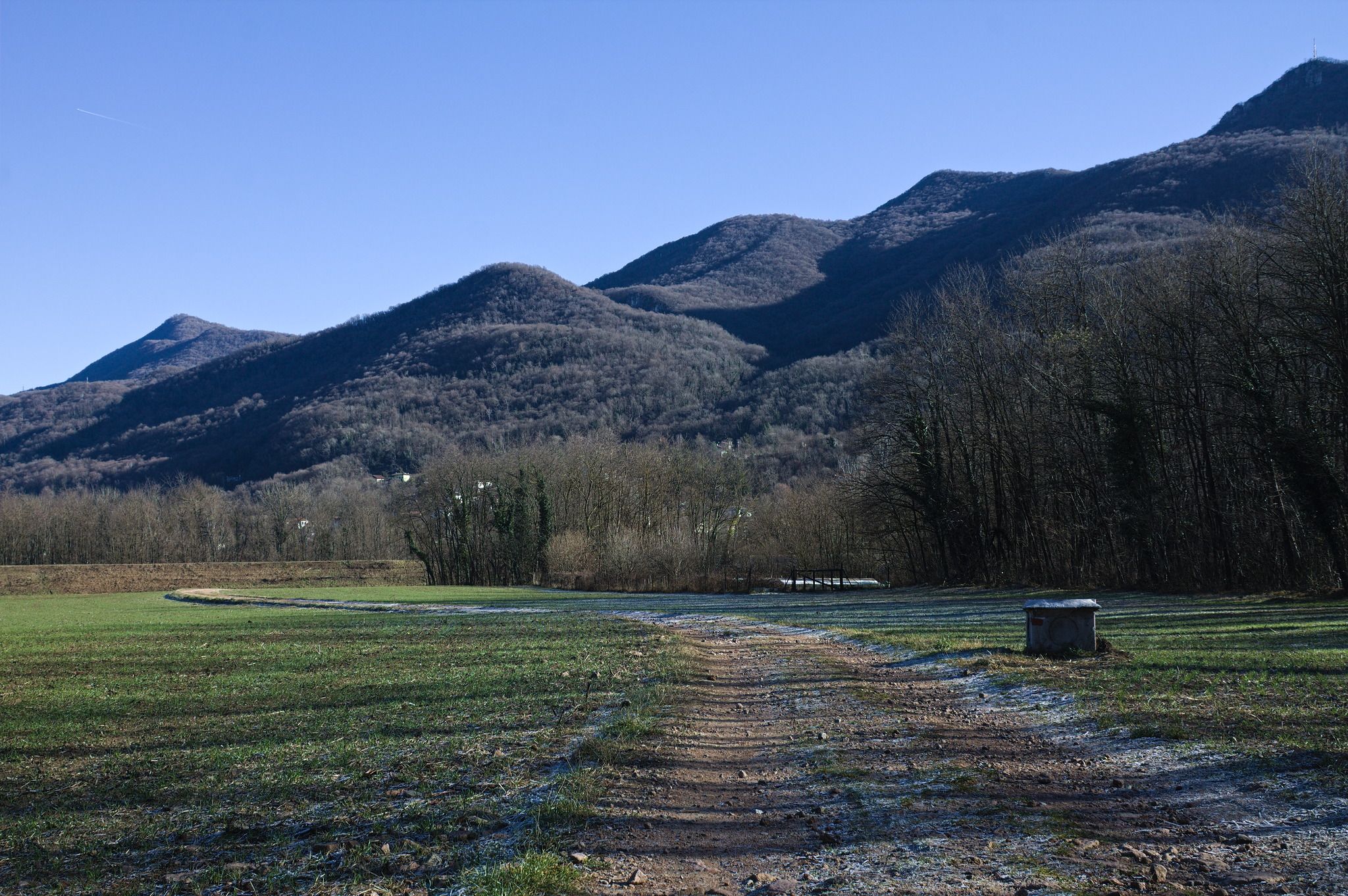 A wide and flat unpaved track passing through a flat grassy area with trees to the sides and rolling hills in the background.