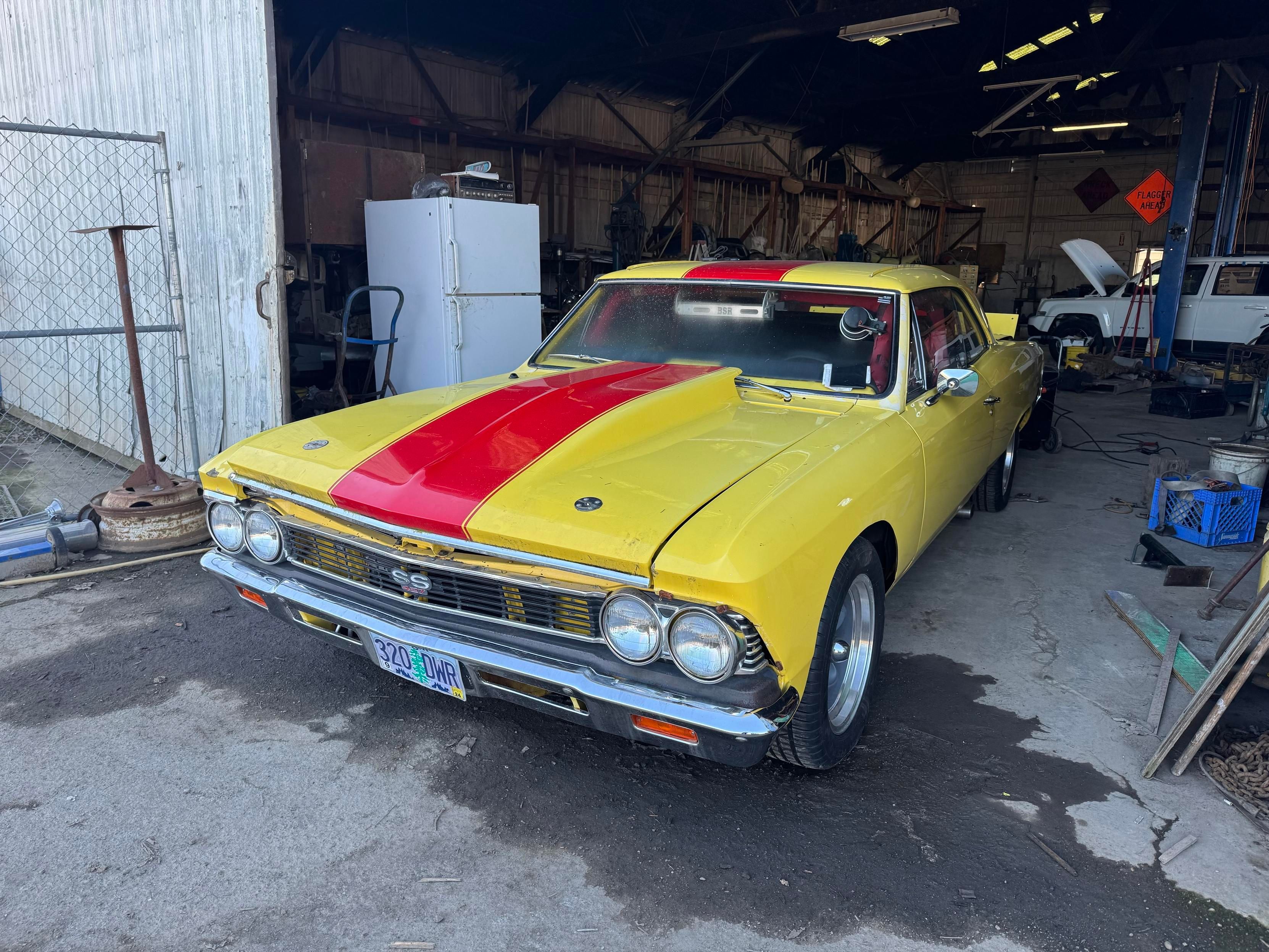 A yellow and red vintage car is parked inside a garage. The car has a classic design, features round headlights, and is labeled as an SS model. The garage contains various tools and equipment, with a refrigerator visible in the background and a few other items 