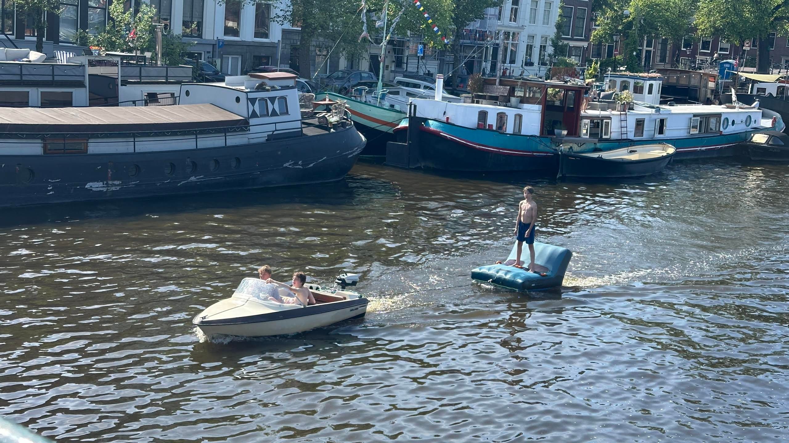 two kids in a tiny speedboat towing an inflatable sofa with another child standing on it