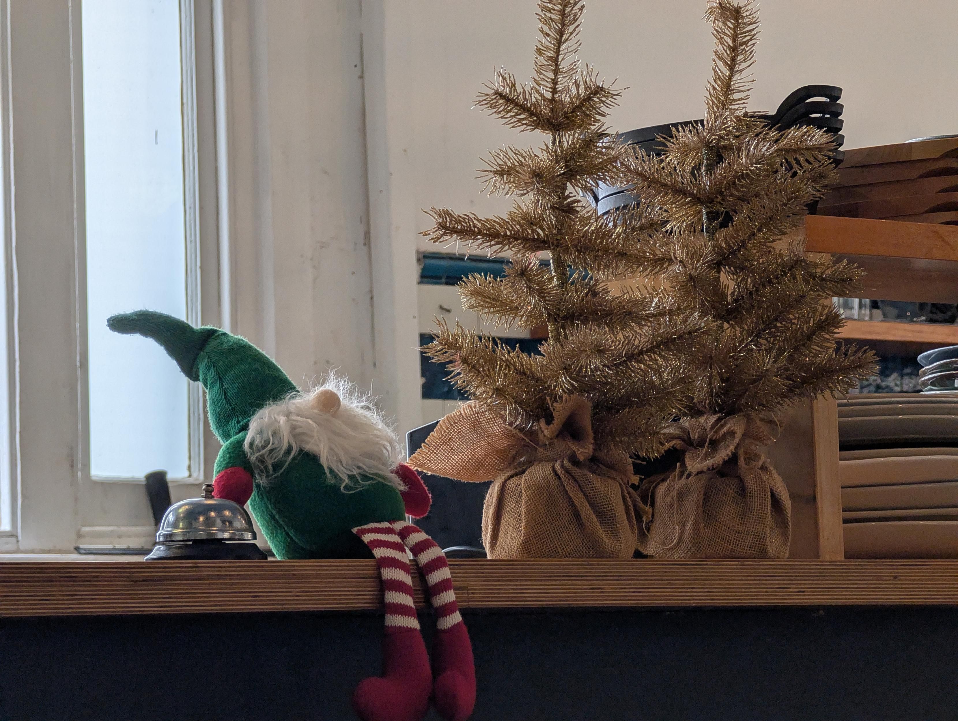 Elf on the bench next to a bell and two dead looking Christmas trees