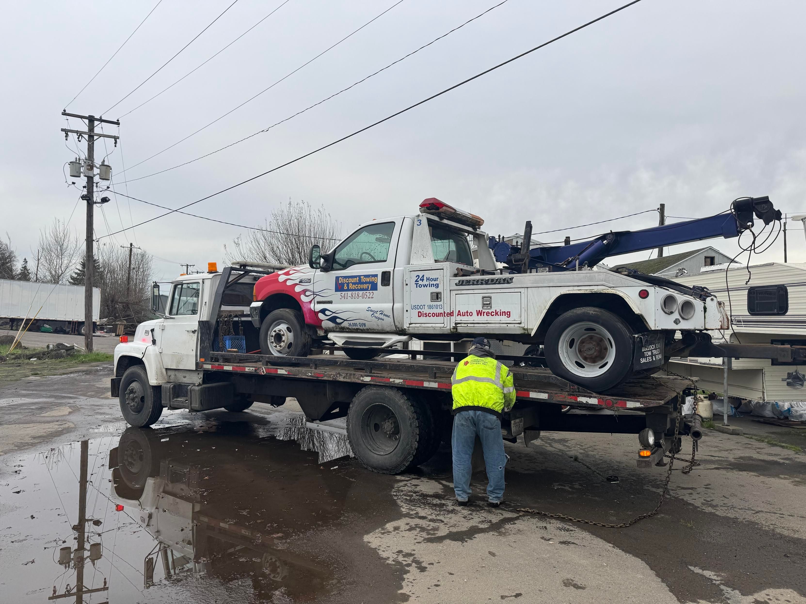 A flatbed tow truck carrying another tow truck is parked in a muddy area, with a man in a bright yellow jacket working on securing the vehicle. Overcast skies and utility poles are visible in the background.
