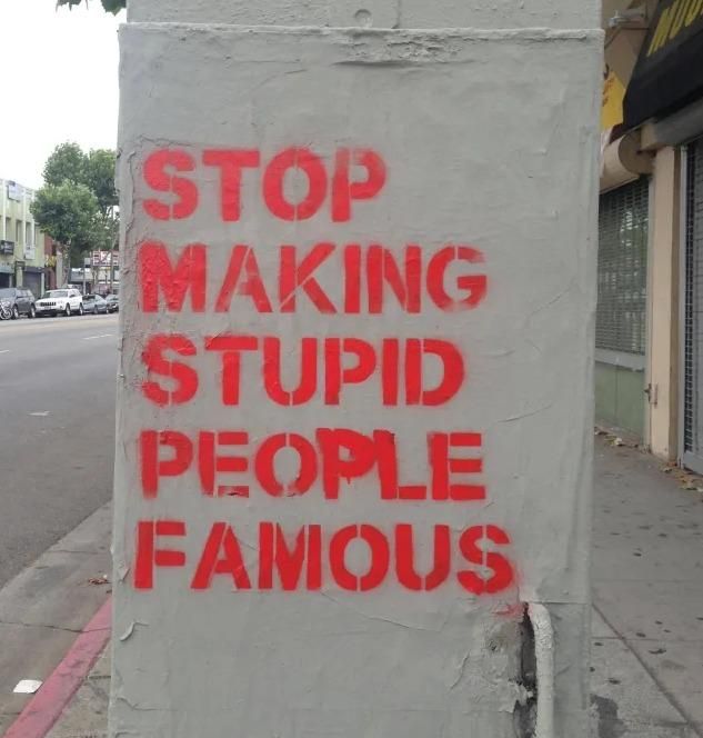 Red stencil graffiti reading “STOP MAKING STUPID PEOPLE FAMOUS” sprayed on a gray utility box along a city sidewalk, with an empty street, parked cars, and storefronts visible in the background. Political and social commentary street art, artist unknown, location unknown.