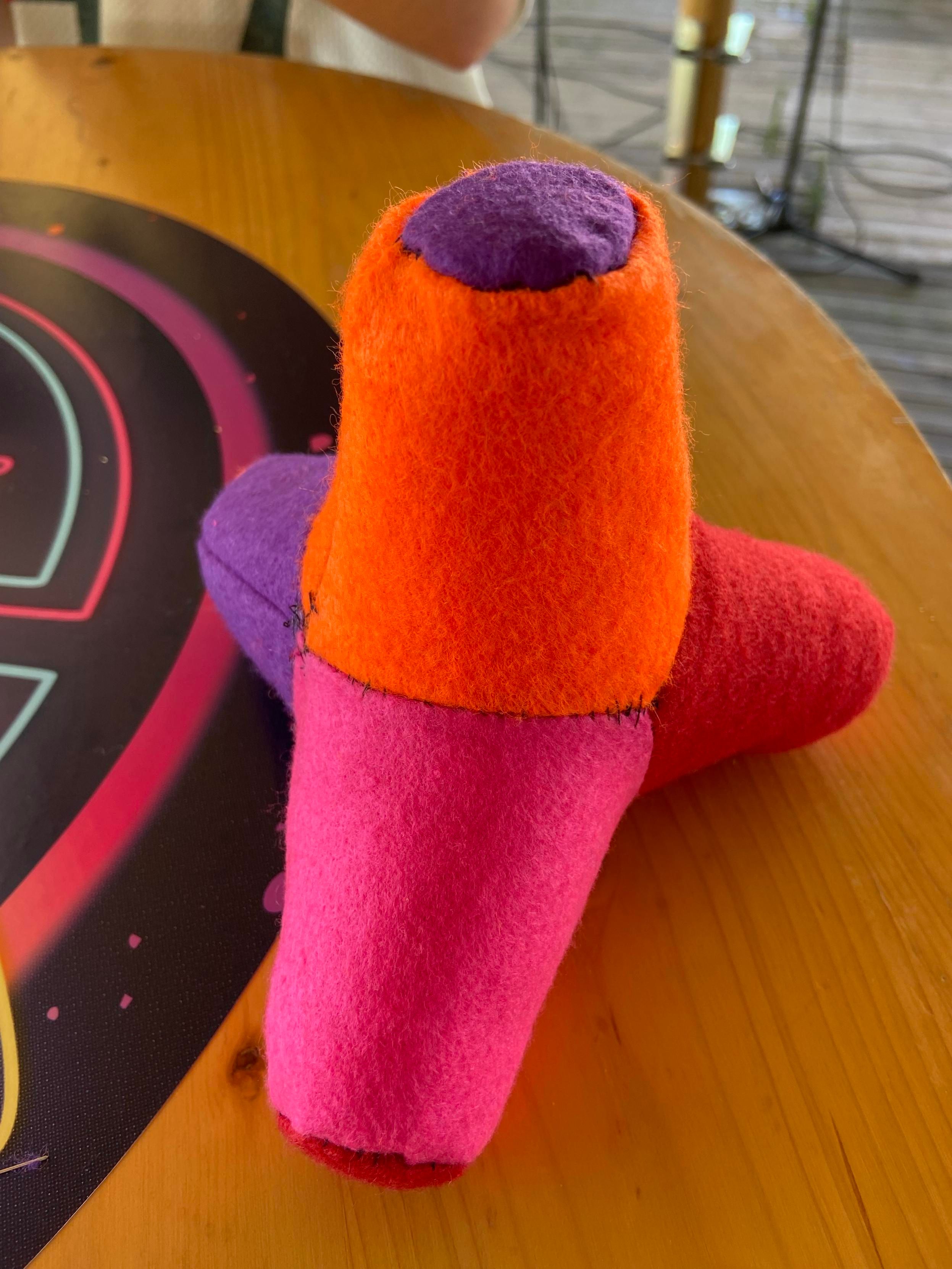 A photo of a plush tetrapod in four colours: orange, pink, purple red. Each leg has a different colour, and the ends of each on another colour.
