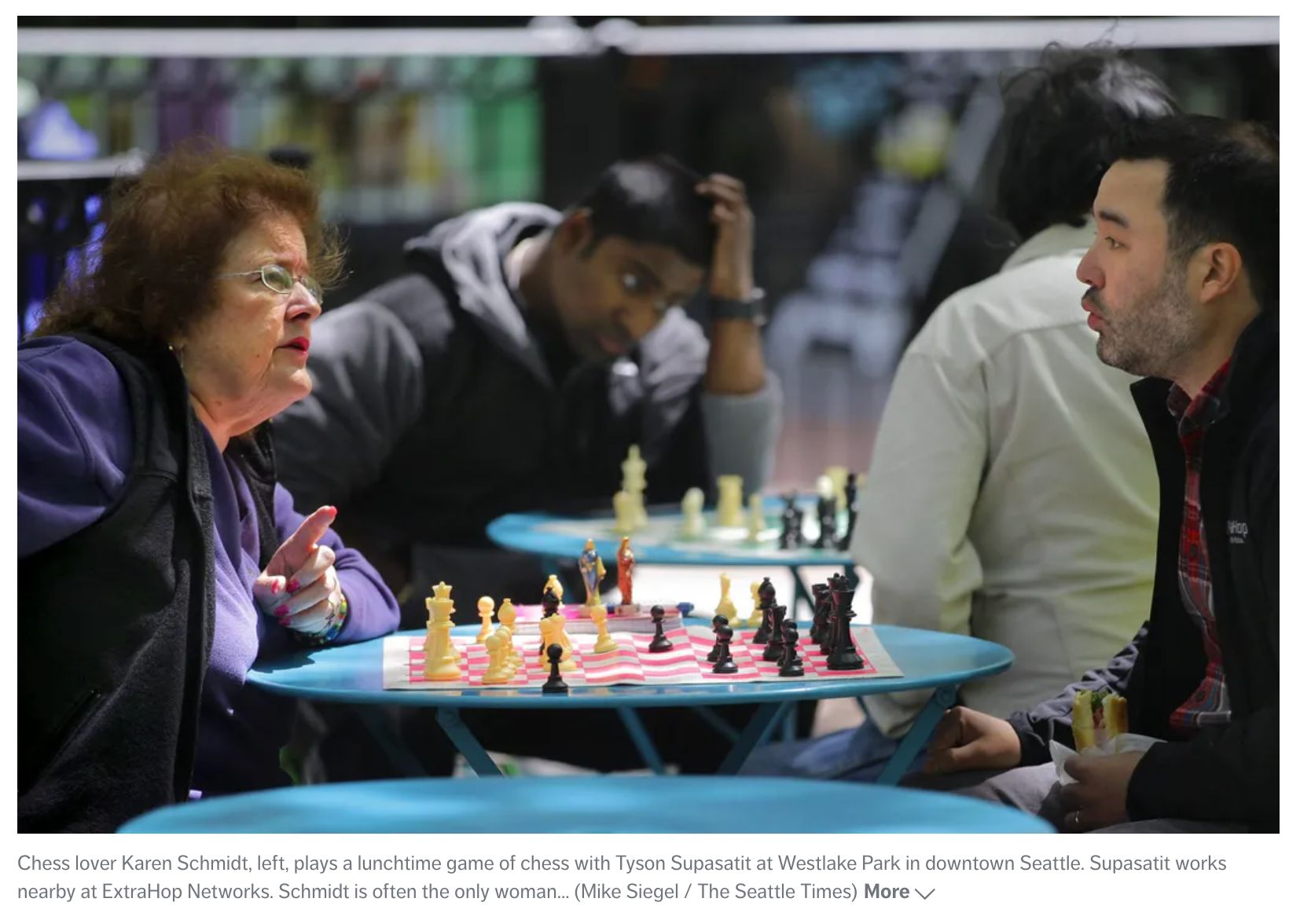 Screenshot from a Seattle Times article on chess in Westlake Park. Photo shows me playing chess with an older lady on my lunch break. In the background, there are two other people playing chess. It's sunny out, and I'm holding a sandwich.