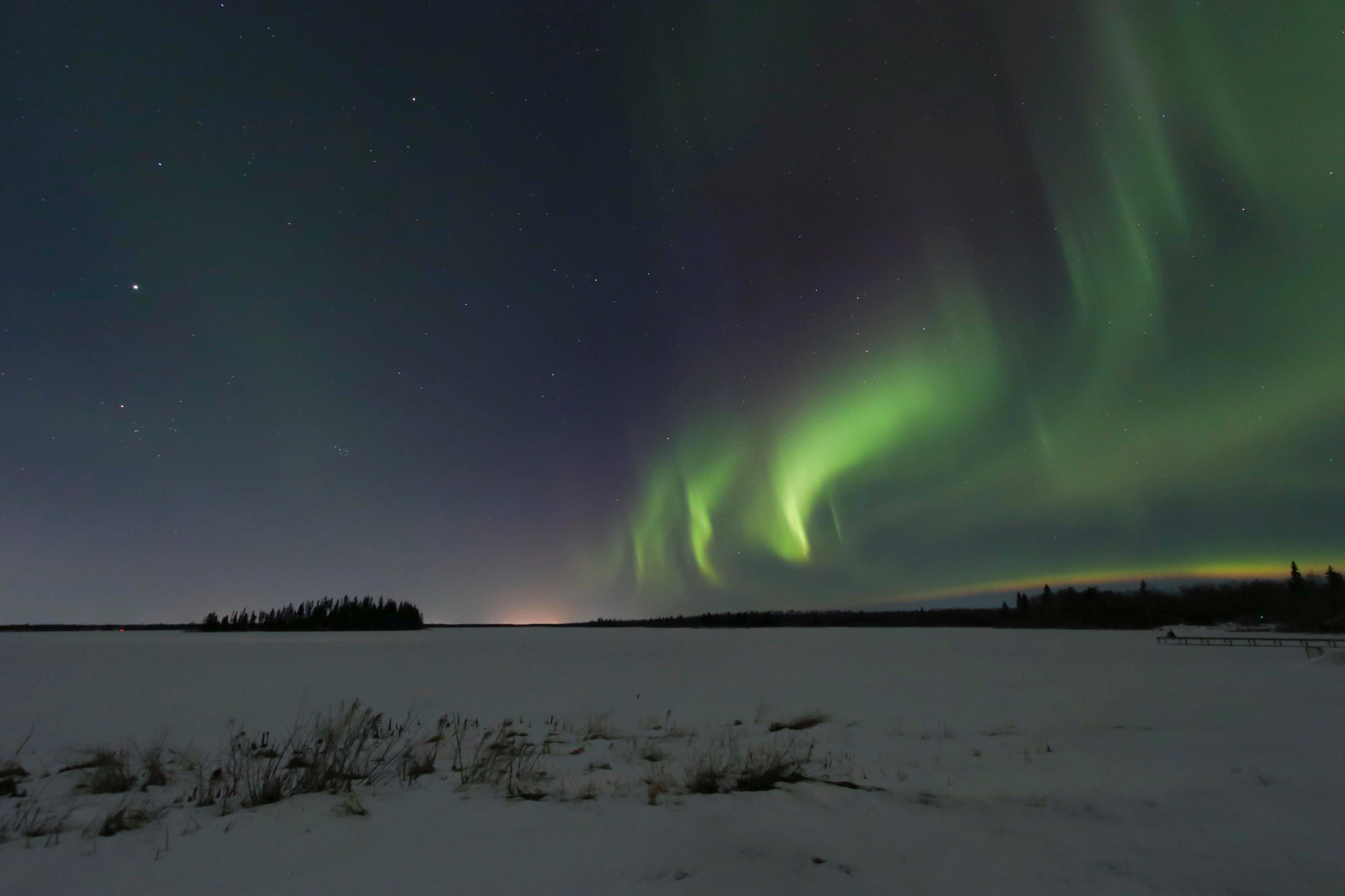 Northern lights glow green with a hint of purple, rippling in the sky above Astotin Lake, at Elk Island National Park in Alberta, Canada. The iconic tree-covered island in the middle of the lake is silhouetted in the background on the left, with the aurora on the right.