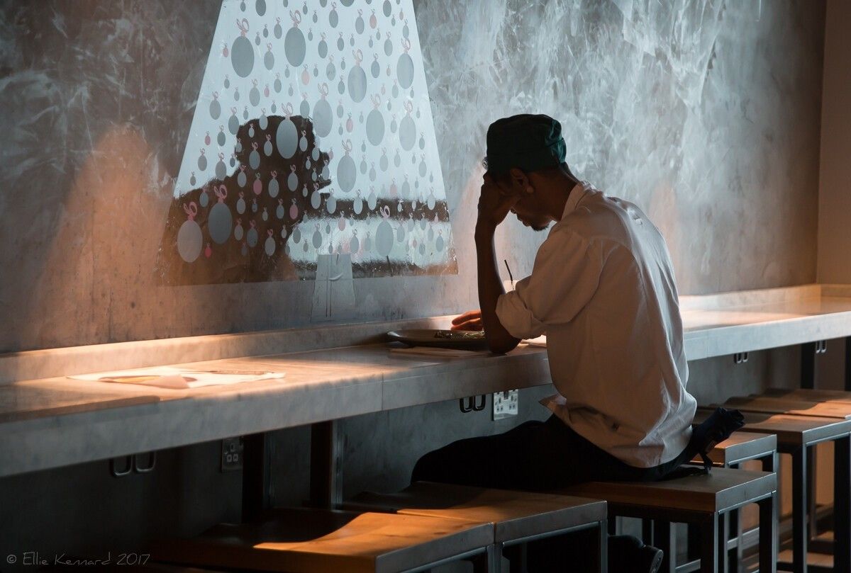 A man is sitting at a long counter with head bowed and one hand to his forehead. There is a patterned wall in front.