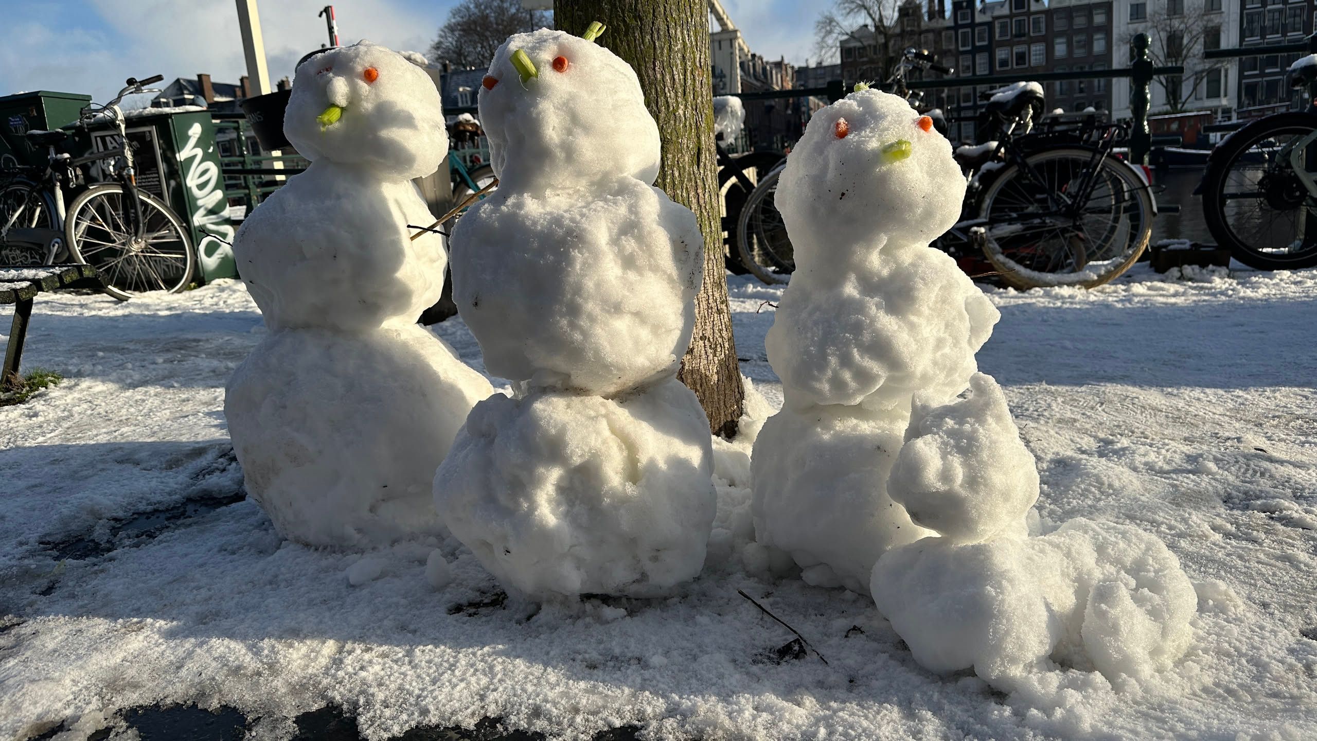 three snow men with a small snow dog. there are bikes and a canal in the background.