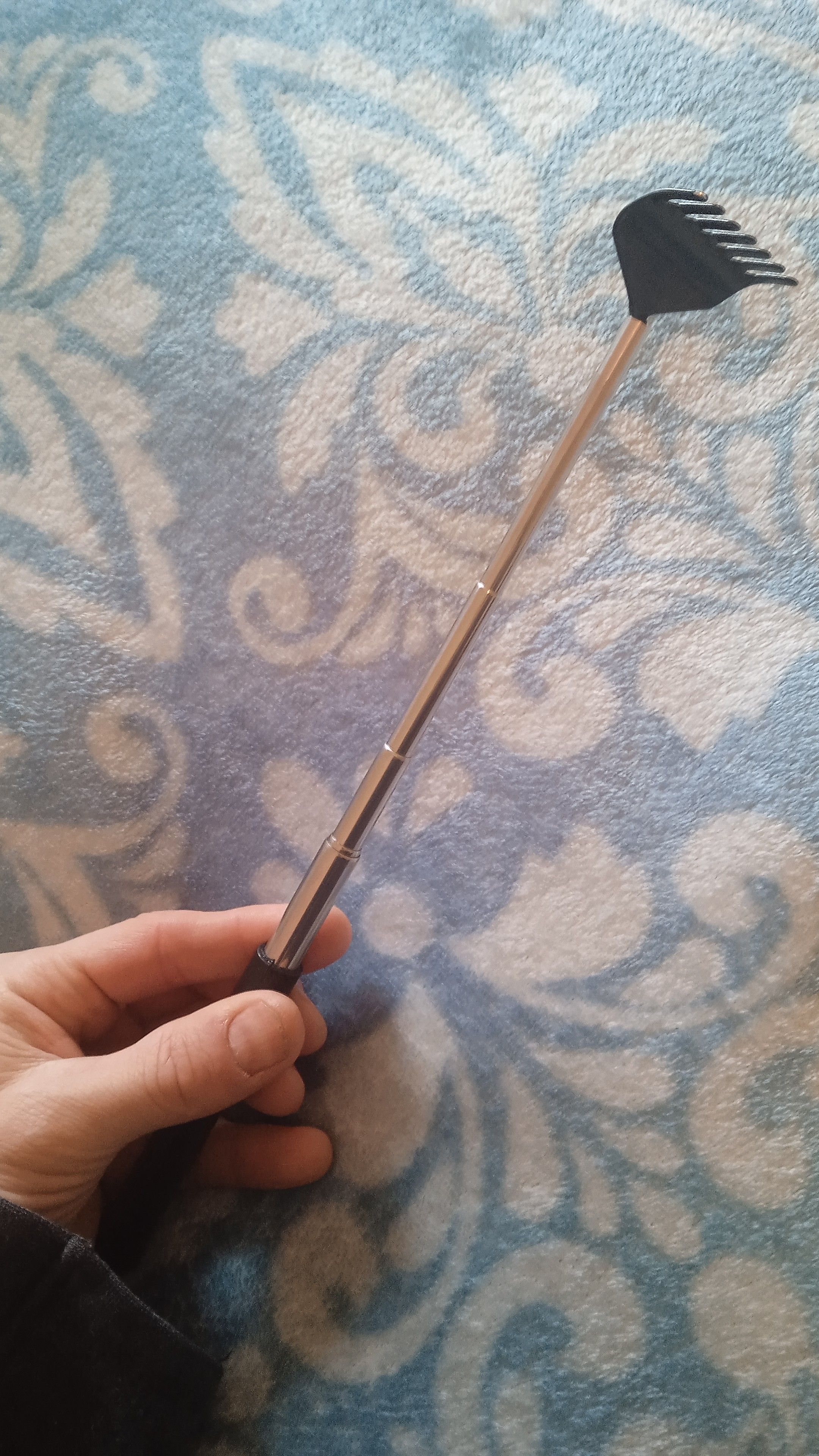 My hand holding a metal back scratcher in the shape of a telescopic rake. On the background, a blue fleece blanket with white decorative motifs