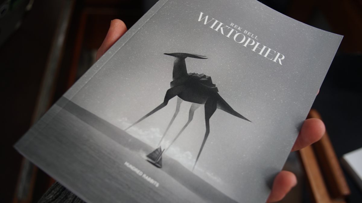 cover of printed copy of Wiktopher, with the cover featuring a sandfin in the desert with a giant long-legged creature.