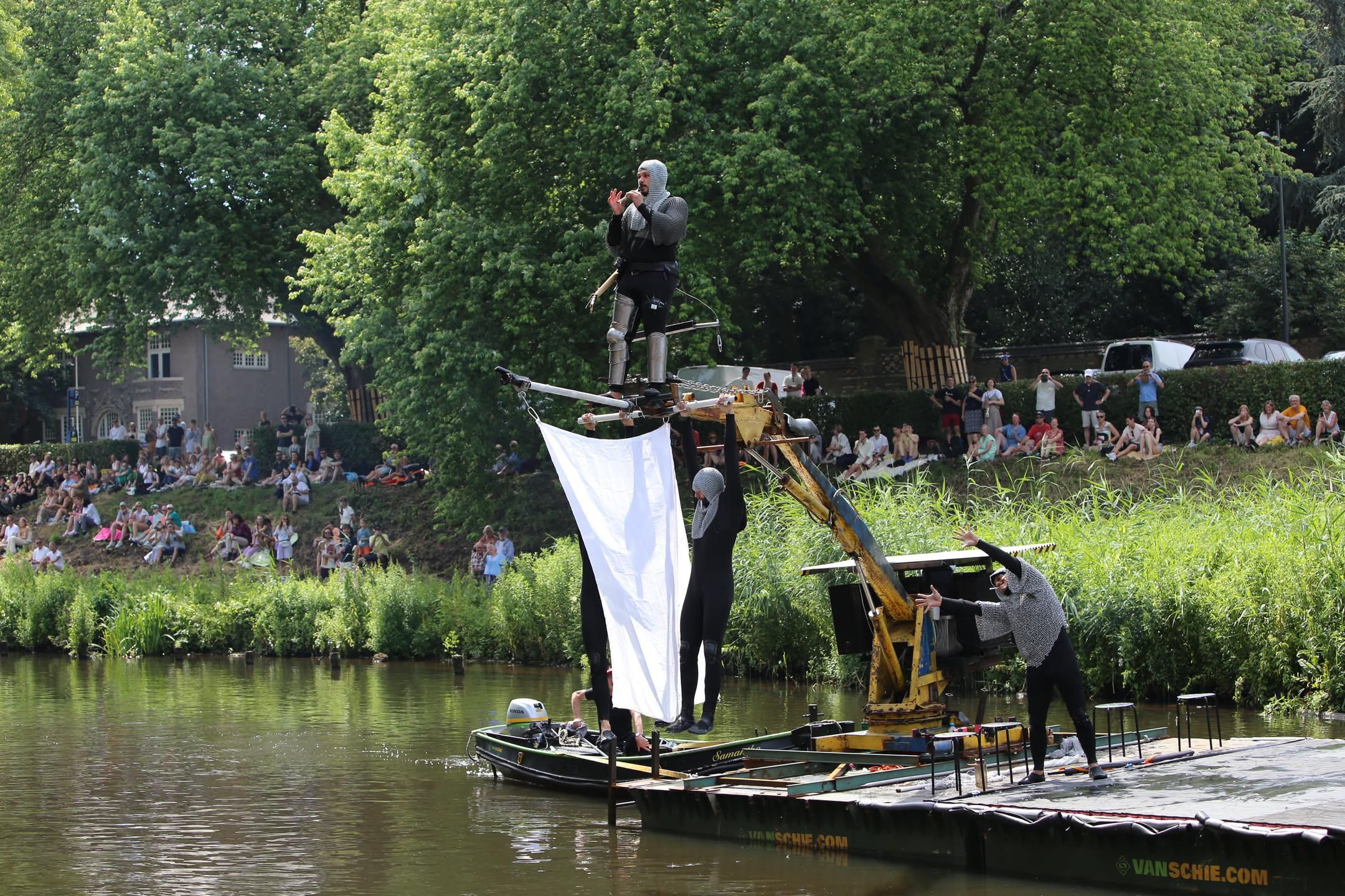 four people wearing chainmail and being lifted by an industrial arm of some sort.  two are hanging from it and one is playing a pipe while standing above it