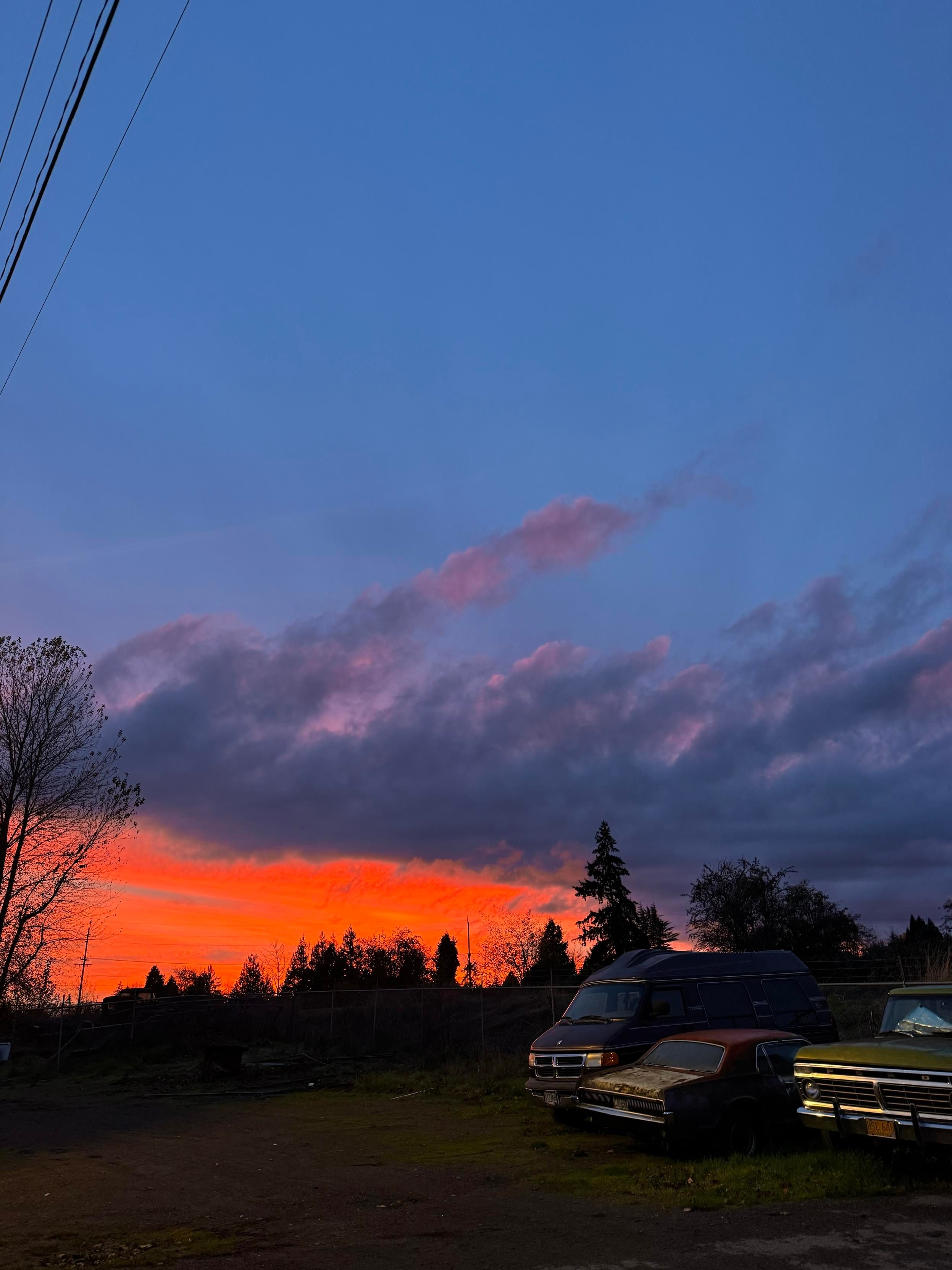 A vibrant sunset displays orange and pink hues against a blue sky, with dark clouds scattered above. Silhouetted trees and power lines are visible in the background, while several parked vehicles, including an old van and cars, are situated in the foreground.