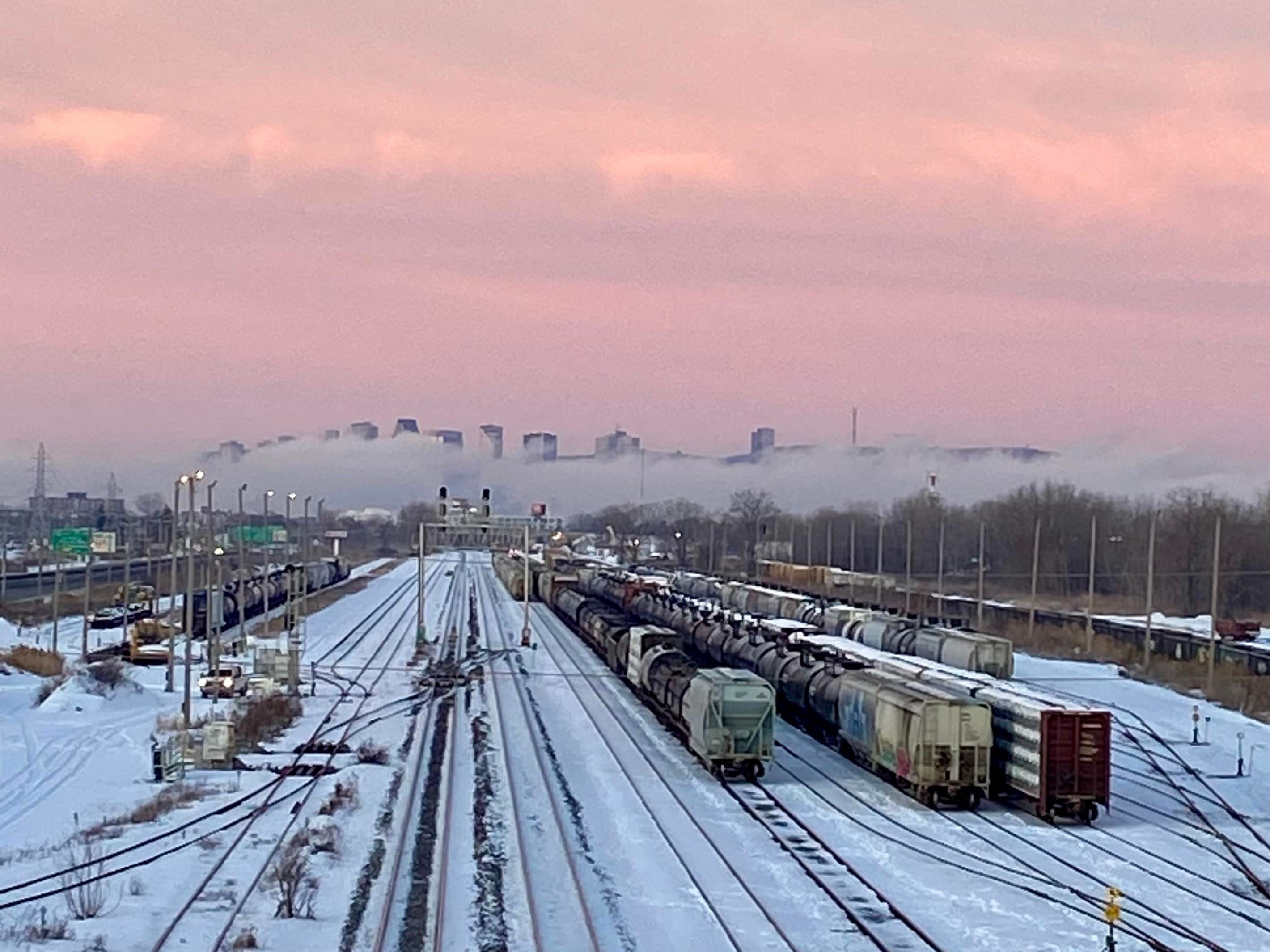 The Montreal skyline seen from a distance, just before sunrise when the sky is pink. It's a cold day and the island is surrounded by mist from the river, with just the tips of the tallest buildings visible. In the foreground is a snowy train yard. 