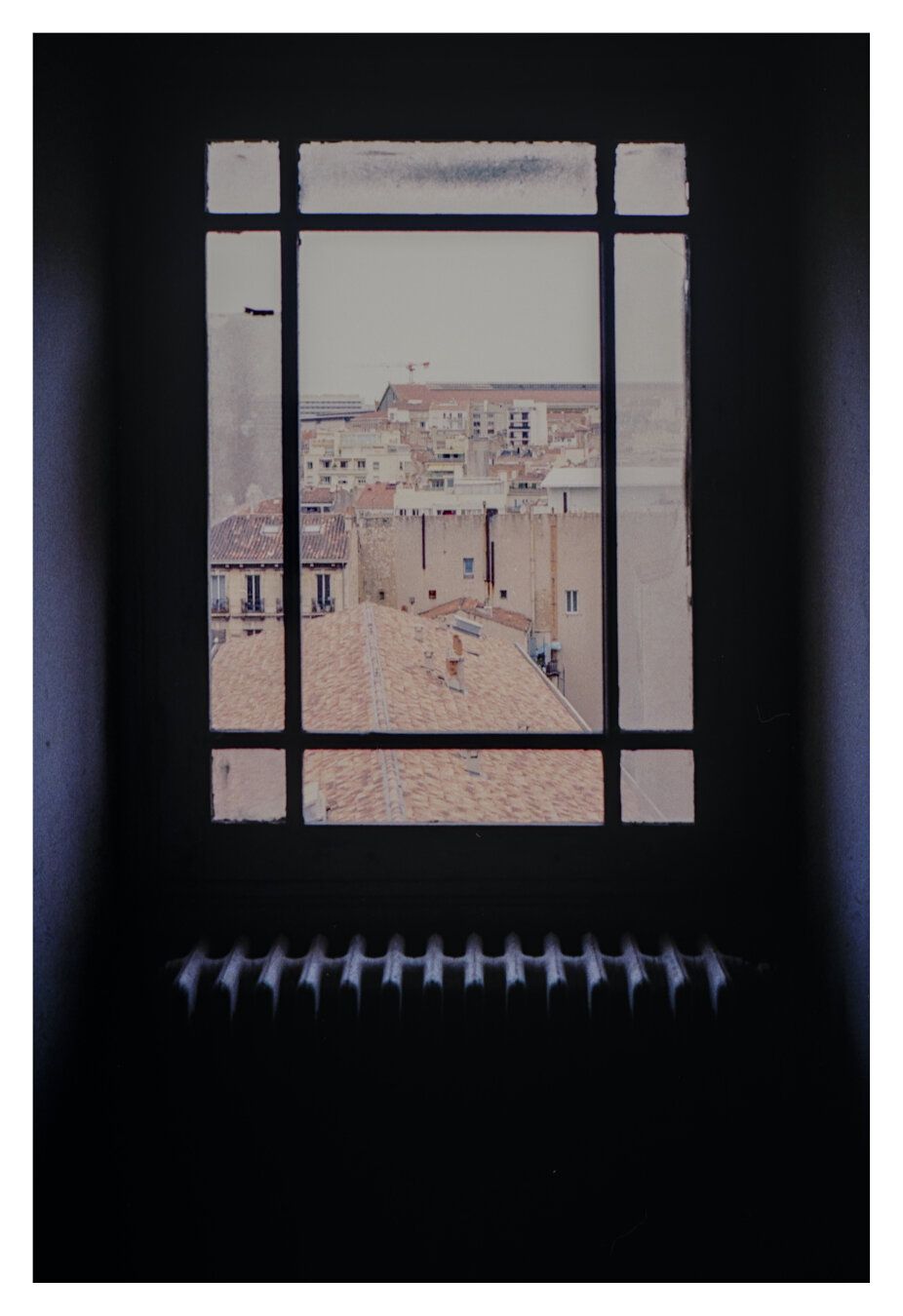 Color photograph of an attic window in the Palais Carli in Marseille. Through the window, you can see the roofs of the surrounding houses.  The window itself is old and dirty. The window frame is almost completely black due to the strong contrast, and below the window, the white surface of the radiator stands out with its striking shape.