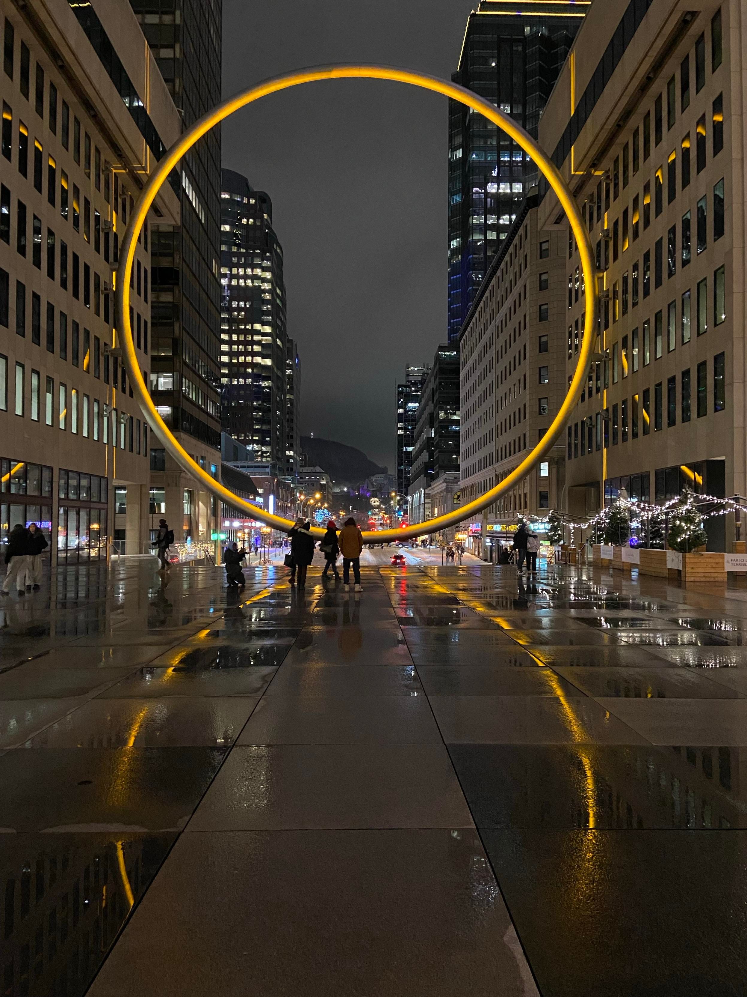 We're standing in downtown montreal at night, looking through the big yellow ring through which you can look up the street and see the McGill campus and arts building. Further behind, we see the cross lit up on Mount Royal. Because we're all about laïcité here, and we don't like to see religious symbols displayed /s 