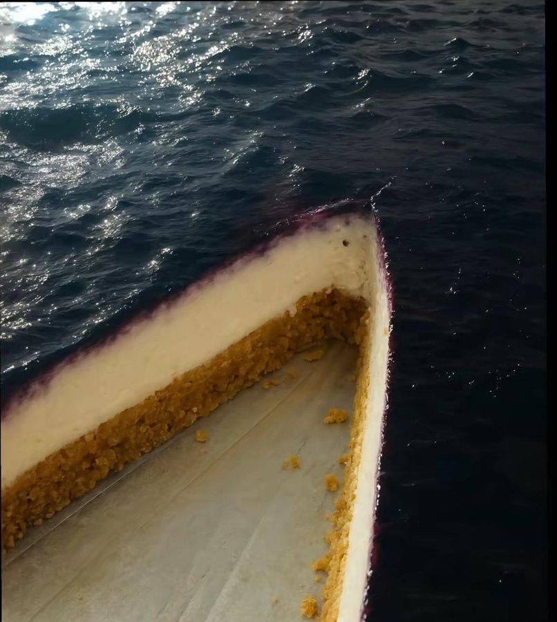 Photo of a missing slice of cheesecake that looks like a cheesecake hull floating in a deep blue ocean. The "ocean" is actually the blueberry topping on the large cheesecake that we don't see completely. 