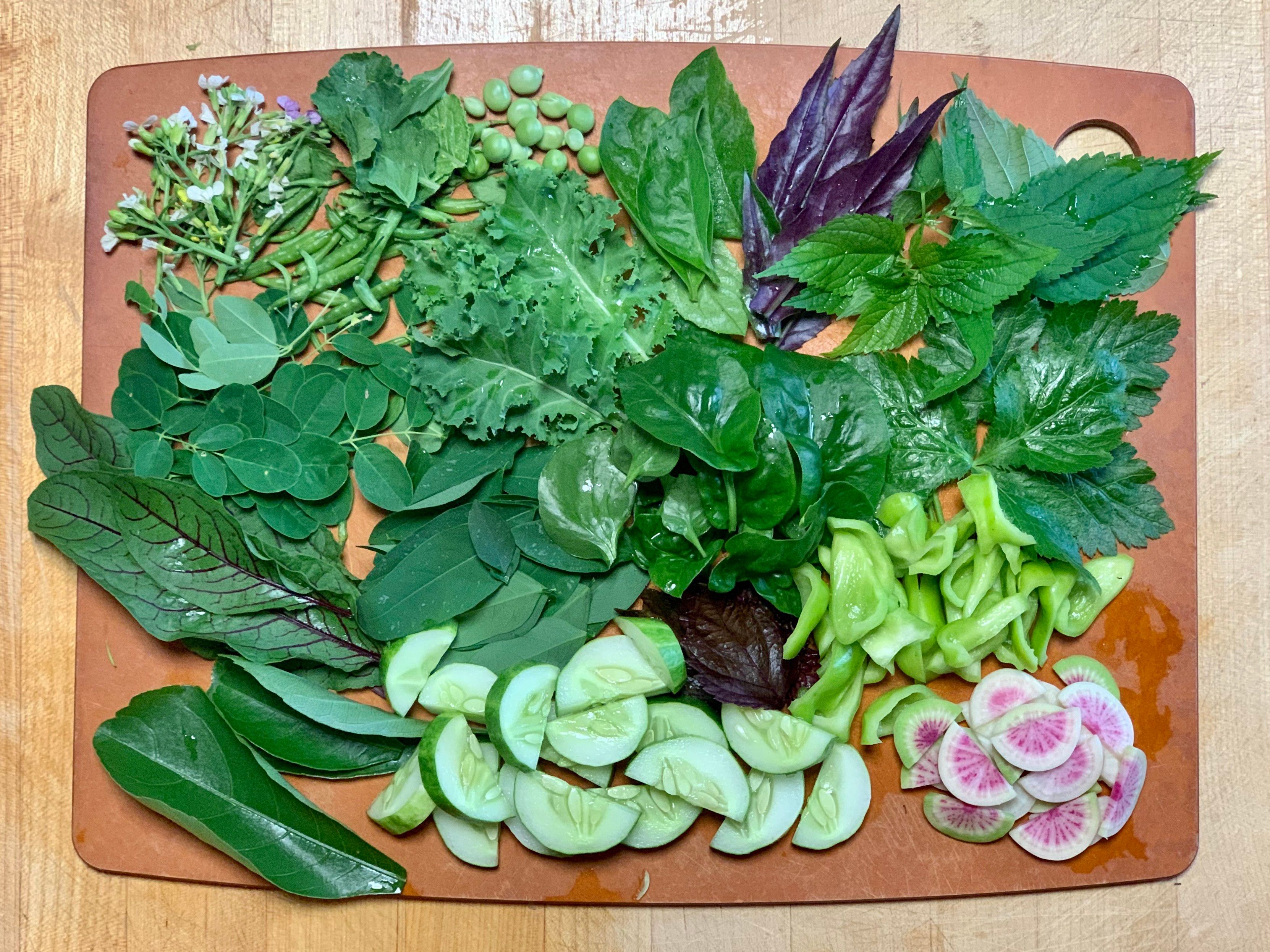 On a wooden cutting board is an assortment of leaves, peas, seed pods, cucumbers, radish, peppers ￼and flowers. 