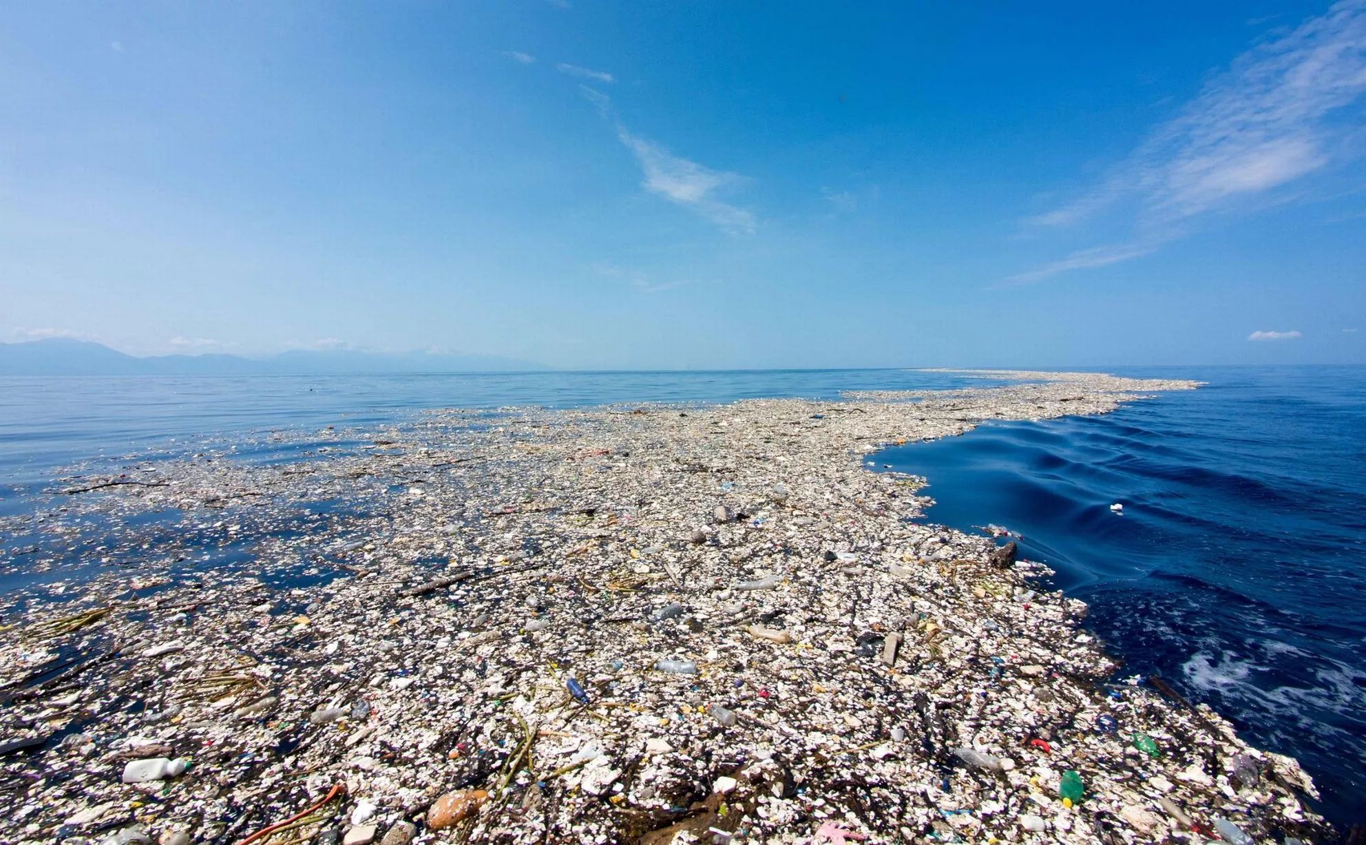 Image shows a lightly clouded but blue sky and an ocean blue with the faint misty hills of a faraway land and a snaking trail of plastic and trash floating on the sea that trails off from the foreground to the horizon.