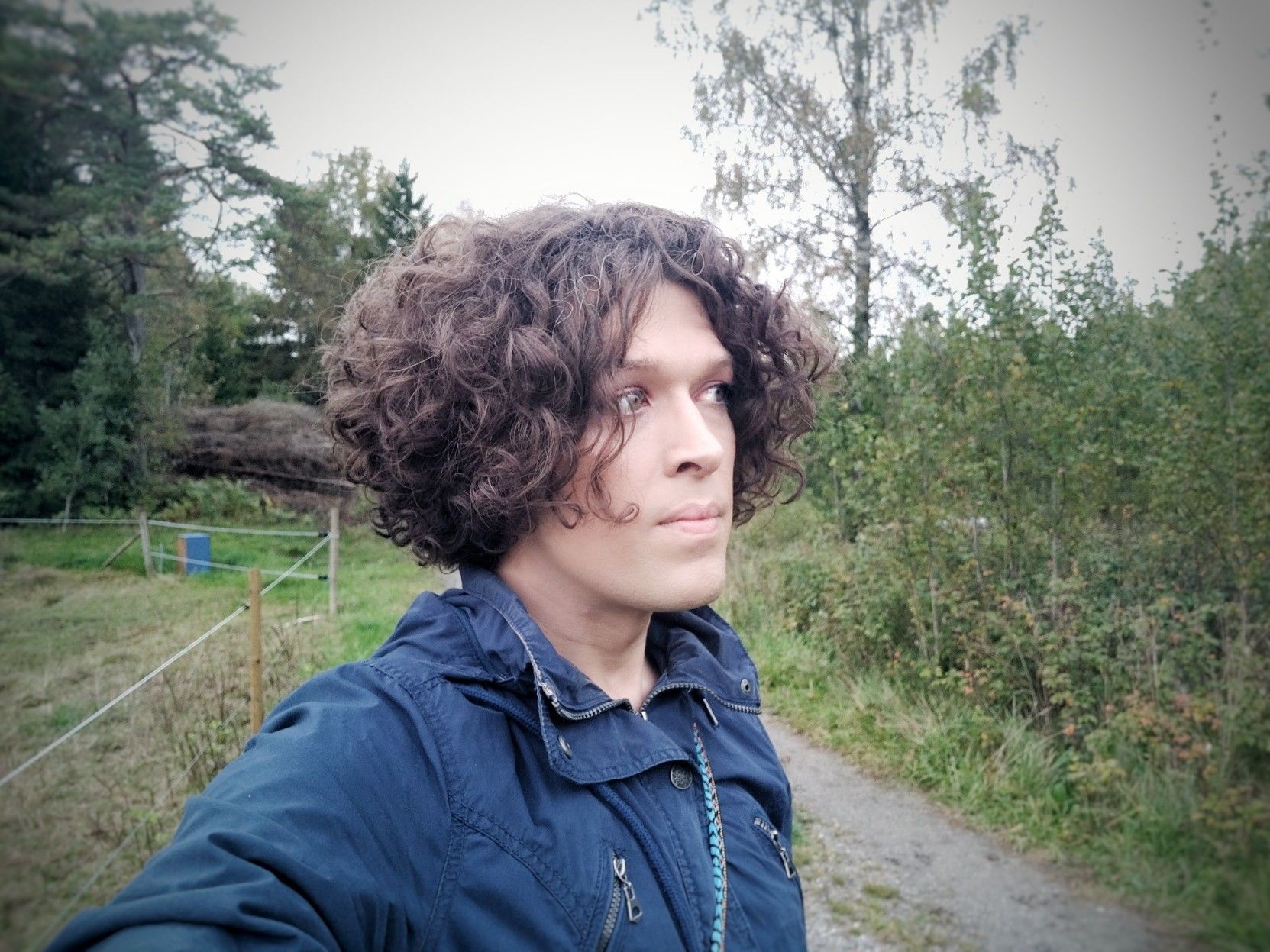 Selfie outdoors in a blue jacket of me with my head turned to your right and looking into the forest.