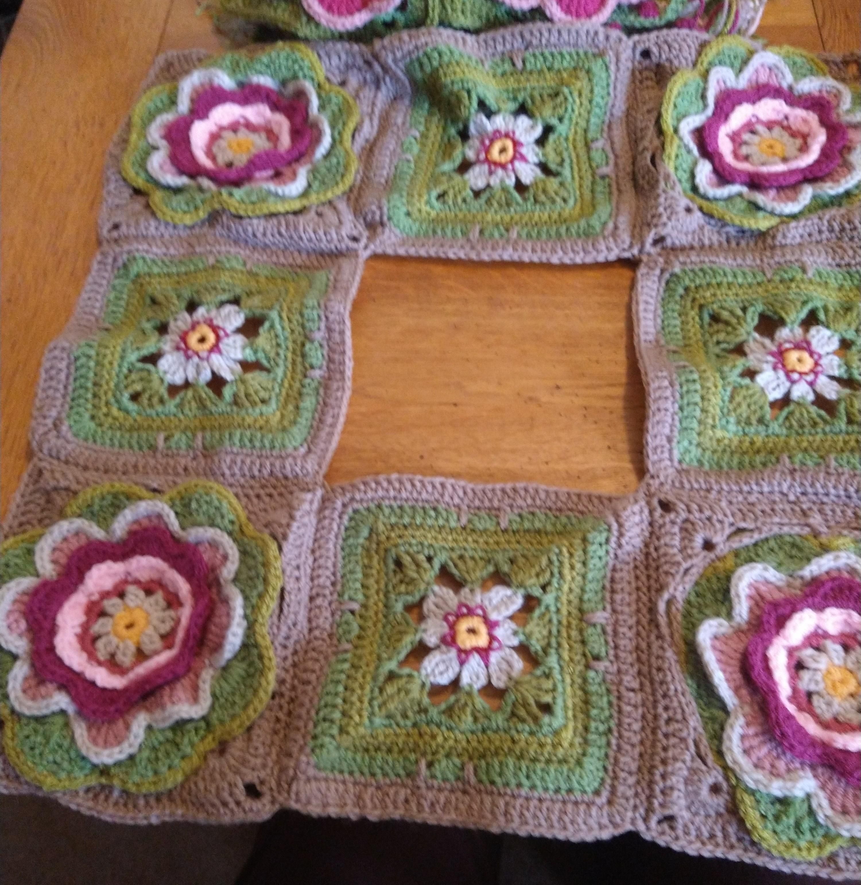 Square crochet blocks in greens and pinks each with a flower motif in pink and green they are pieced together to form a square with a central square missing 